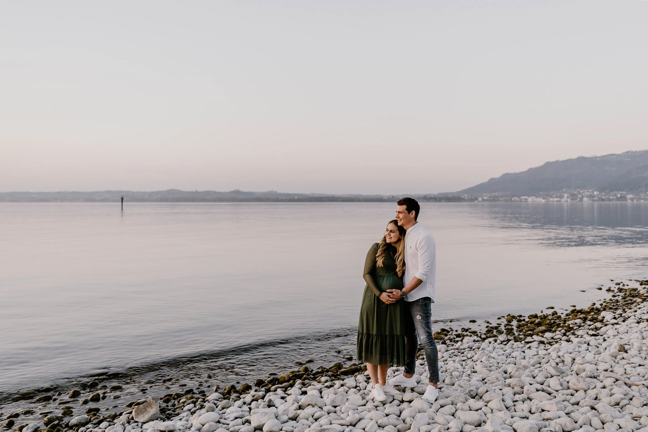 Ein Paar steht am Strand mit Steinen, umgeben von Wasser und Bergen im Hintergrund, bei Sonnenuntergang.