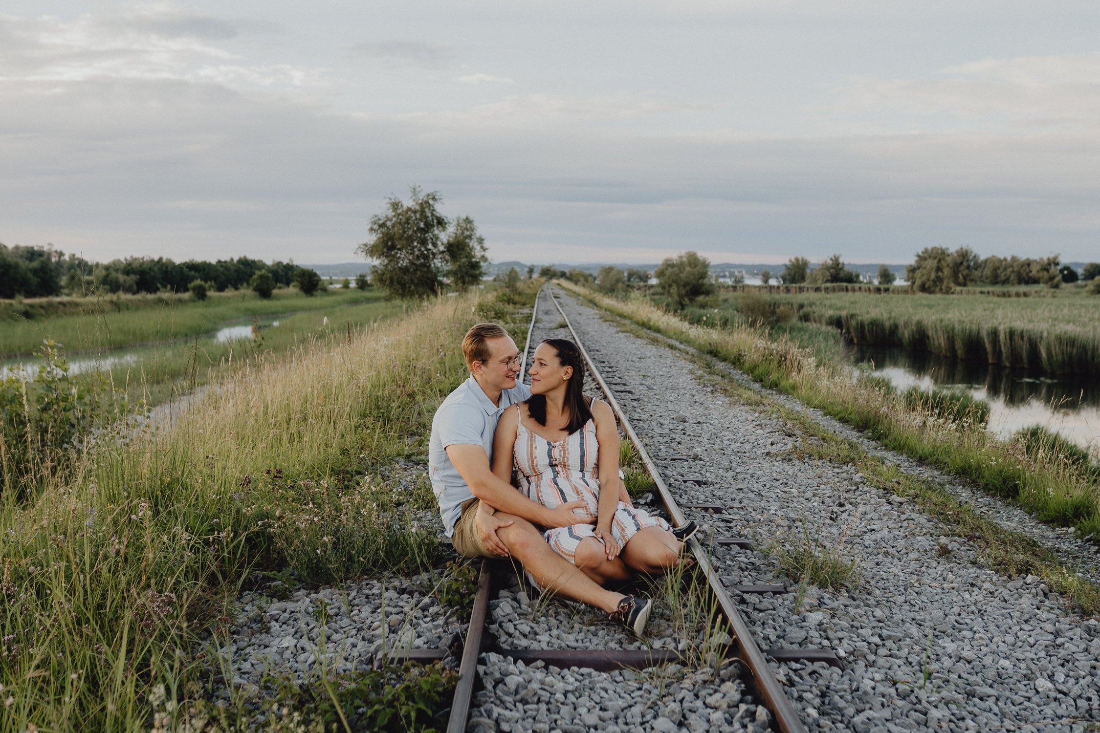 Ein junges Paar sitzt auf Eisenbahnschienen in einer ländlichen Umgebung mit Fluss, Wiesen und Bäumen, umgeben von Natur, während sie sich liebevoll anschauen.