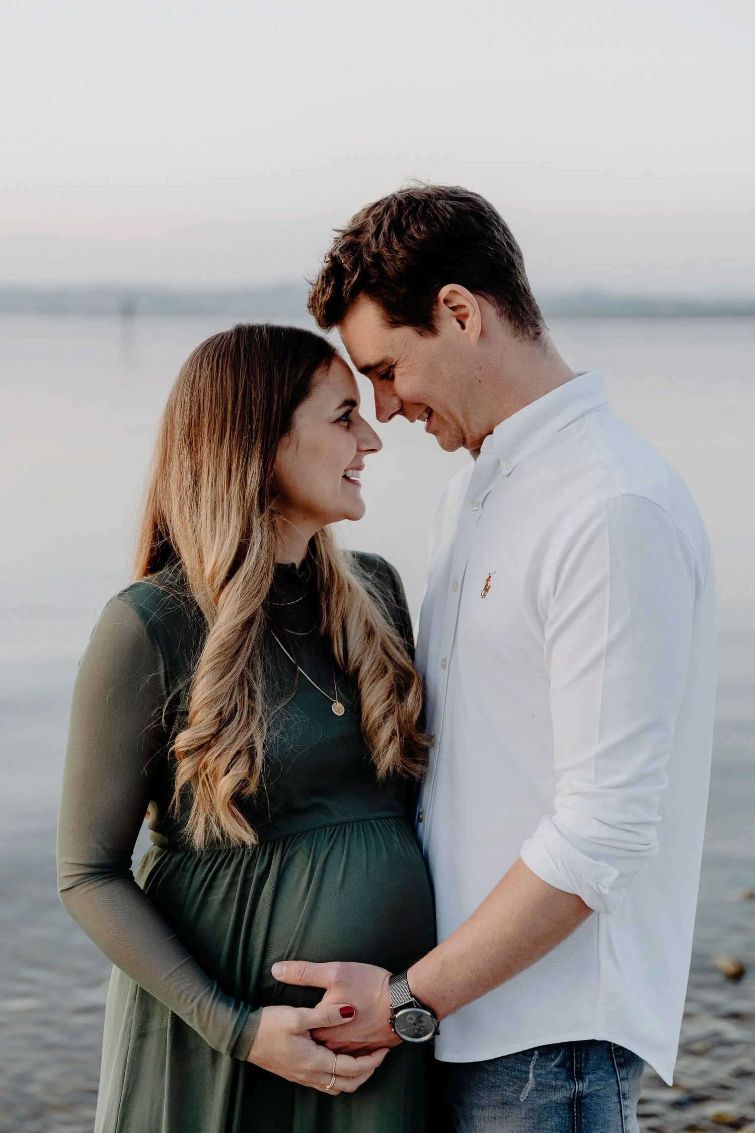 Ein schwangeres Paar steht am Wasser, lächelt sich an, mit ihren Stirn aneinander und hält die Hände auf den Babybauch.