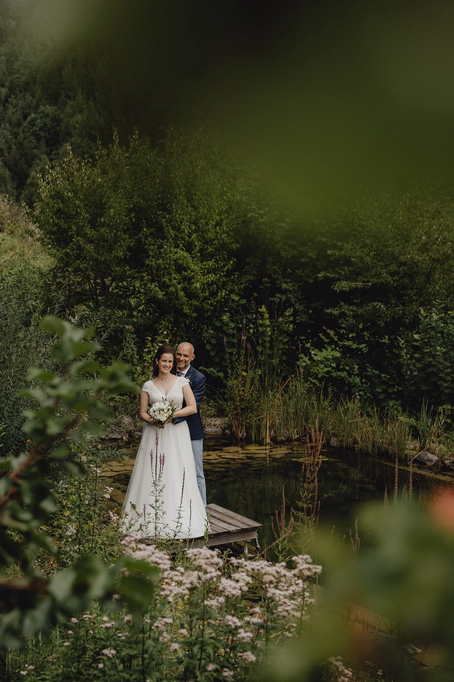 Ein Brautpaar steht auf einer kleinen Holzplattform neben einem Teich, umgeben von grünen Pflanzen und Blumen, bei ihrem Hochzeitsshooting im Freien.
