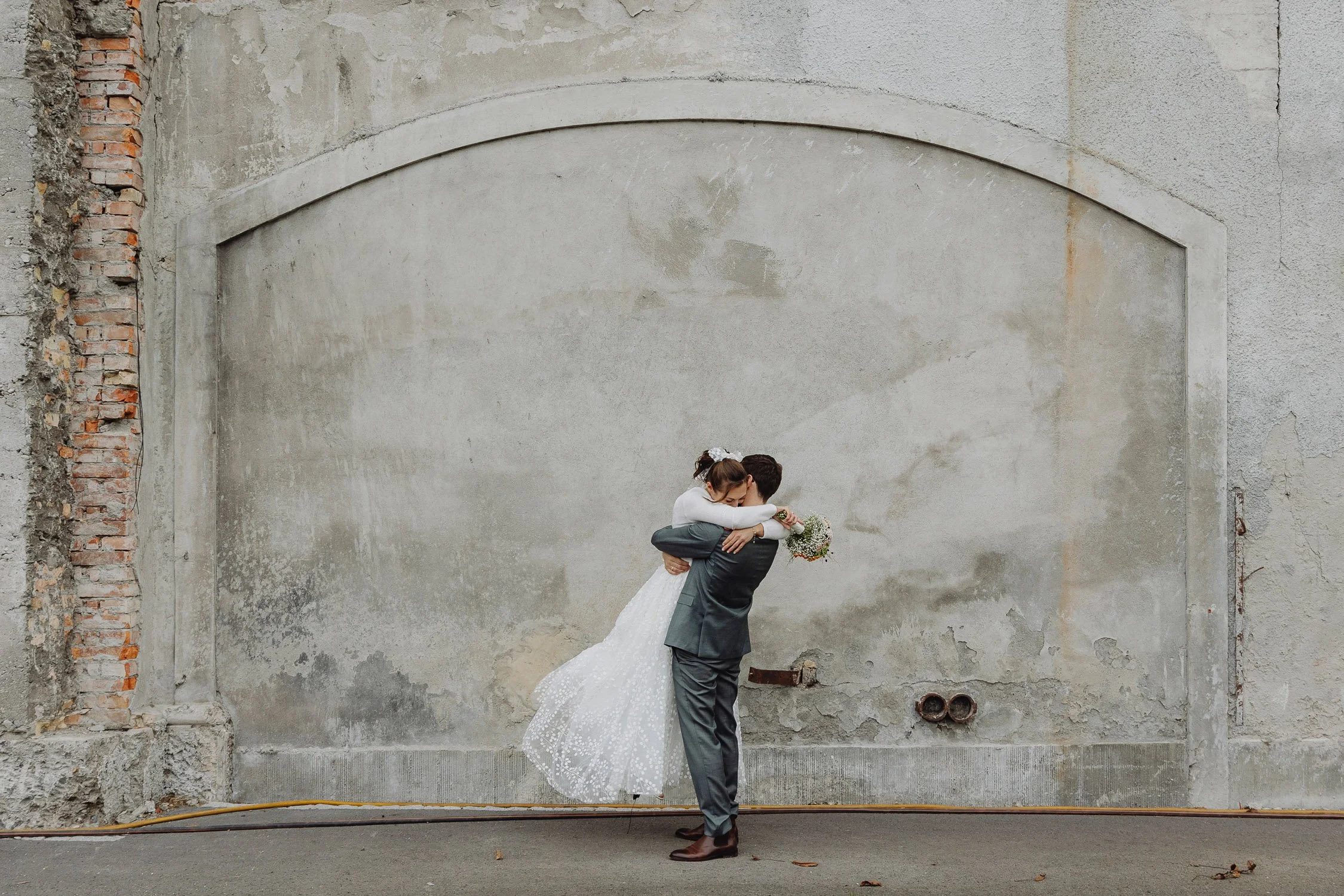 Ein Brautpaar umarmt sich vor einer grauen, ungepflegten Wand, die an einer alten, teils beschädigten Mauer steht, die teilweise aus Ziegelsteinen besteht. Der Boden ist asphaltiert, einige Blätter liegen auf dem Boden.