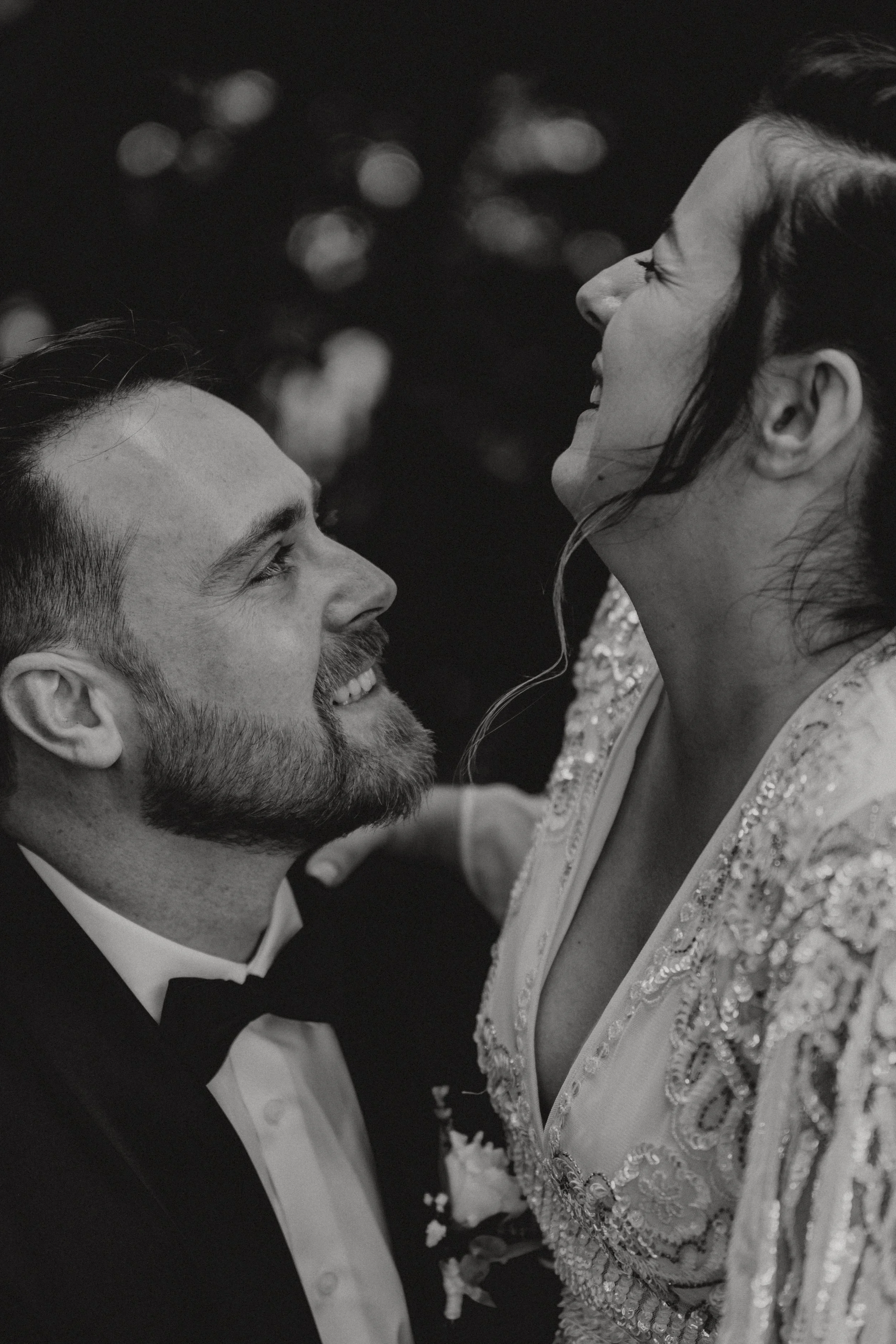 Ein lachendes Paar bei einer Hochzeit, der Mann trägt einen schwarzen Anzug mit Fliege, die Frau trägt ein gesticktes Kleid.