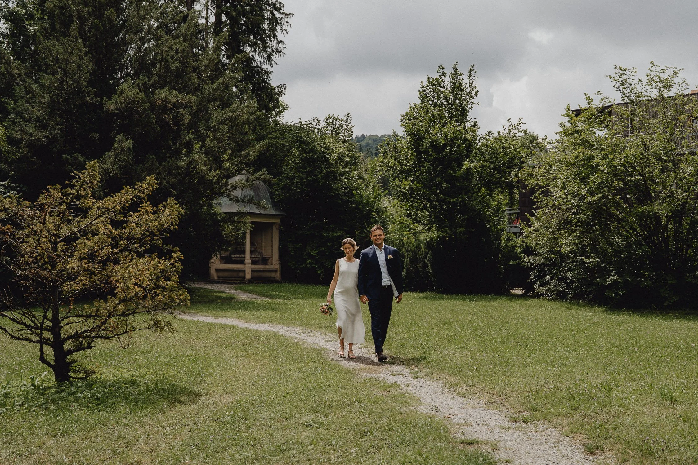 Ein Brautpaar läuft Hand in Hand auf einem grünen Rasenweg, umgeben von Bäumen und einer Parklandschaft, bei bewölktem Himmel.
