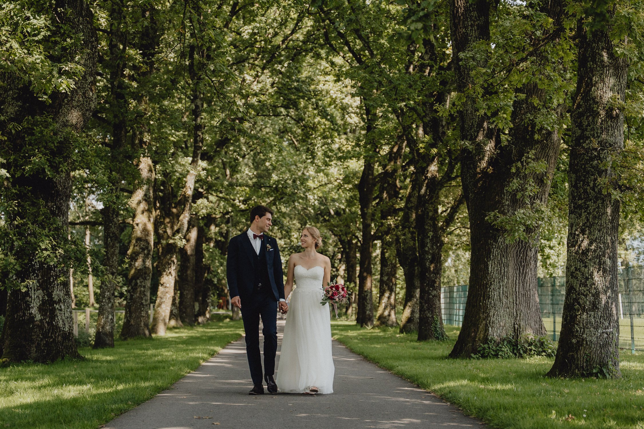 Ein Brautpaar spaziert Hand in Hand durch einen grünen Park mit großen Bäumen an einem sonnigen Tag.