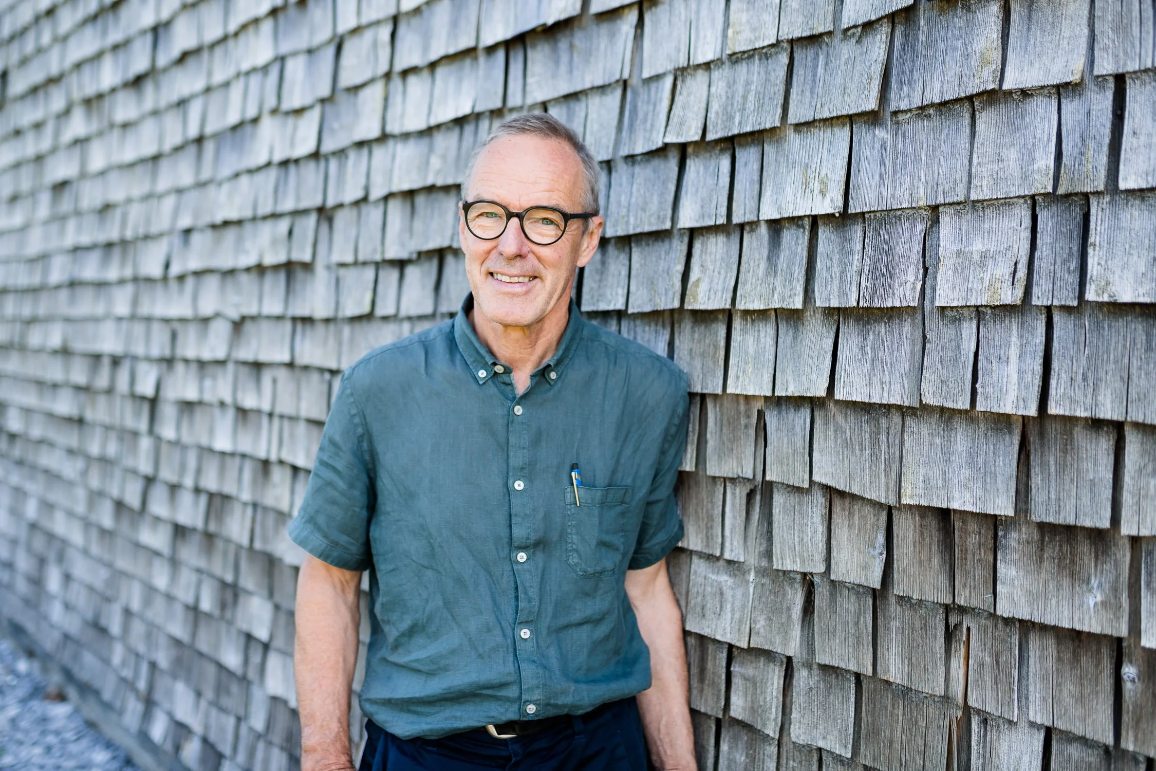 Älterer Mann mit Brille trägt blaue Hemd und lächelt, steht an einer Holzschindeltapete.