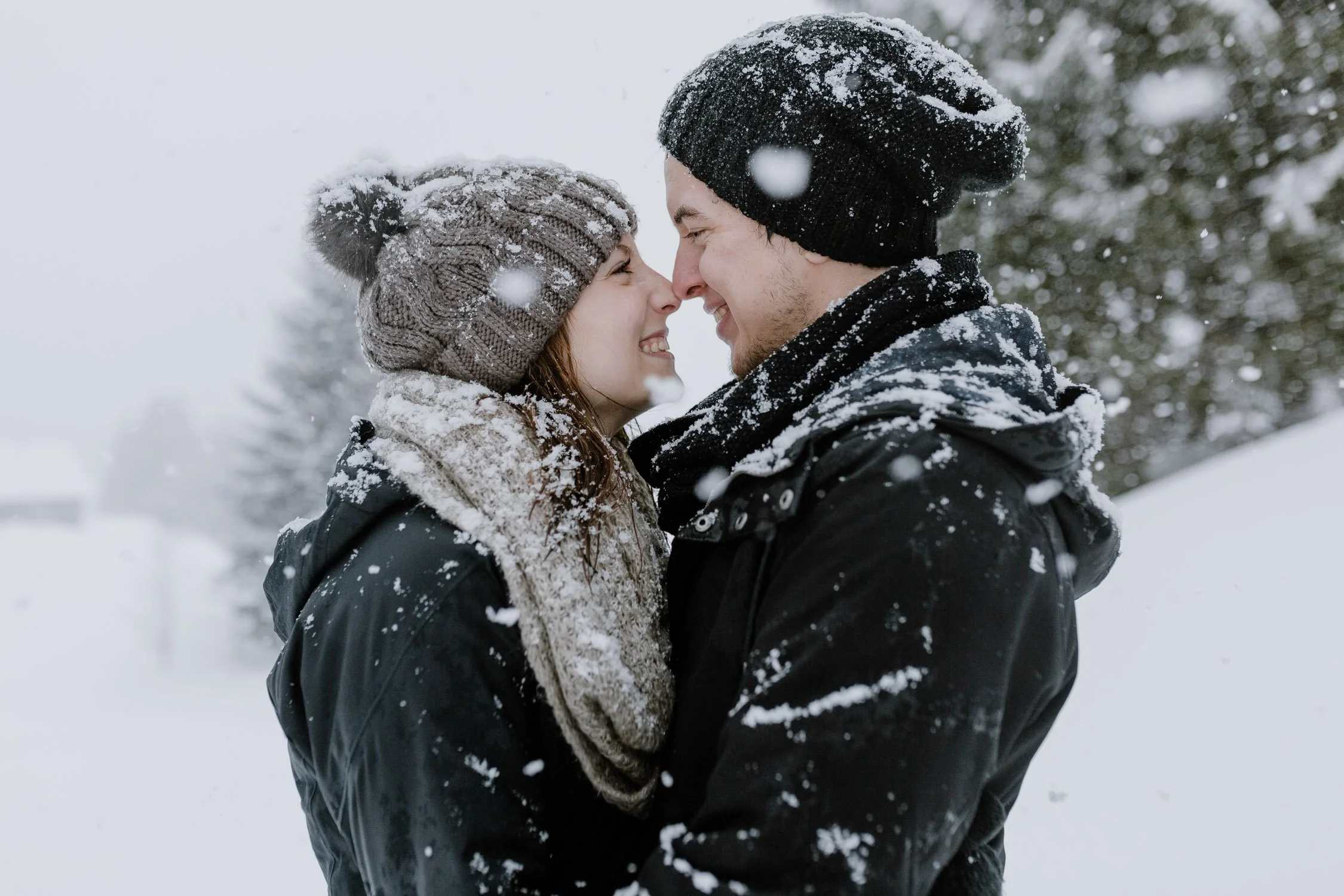 Ein junges Paar steht sich in Schneeland gegenüber, beide tragen Winterkleidung und Mützen, während Schneeflocken fallen.