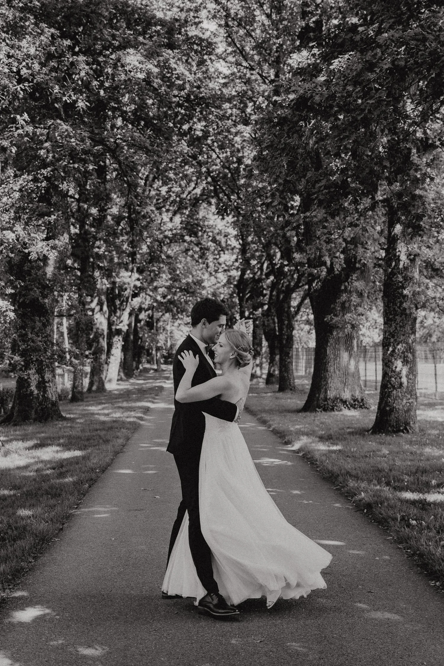 Ein brautpaar tanzt auf einem Weg in einem Park mit großen Bäumen.