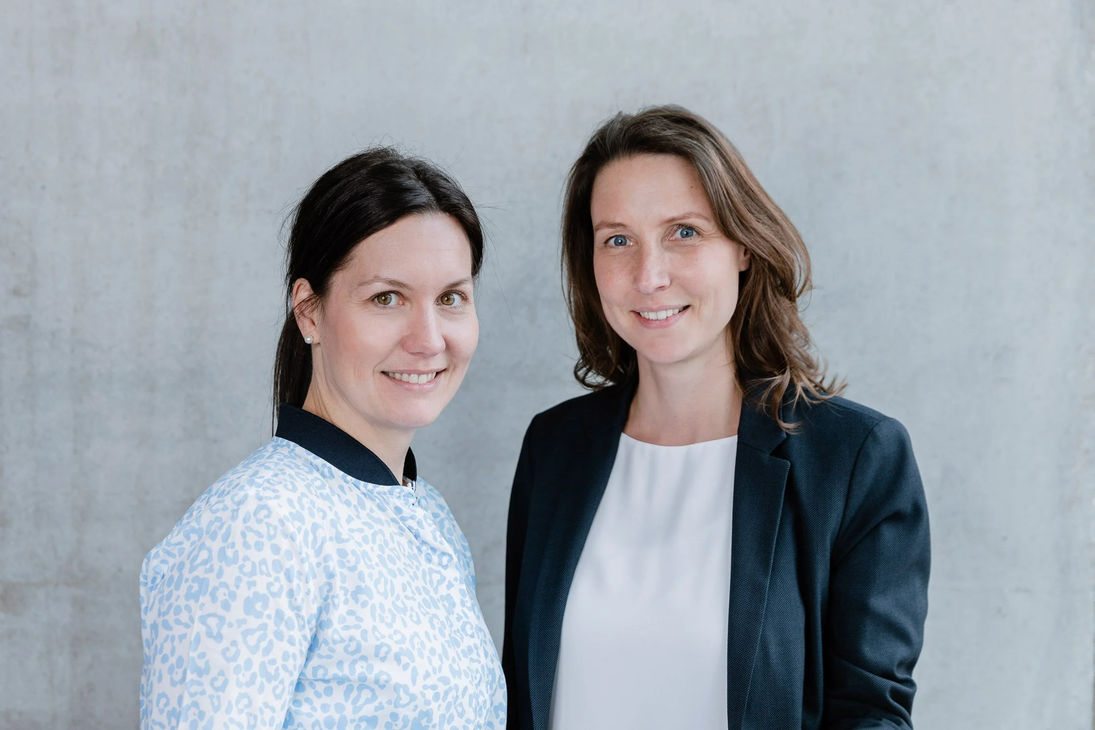 Zwei Frauen stehen nebeneinander vor einer neutralen Wand, lachen und schauen in die Kamera.
