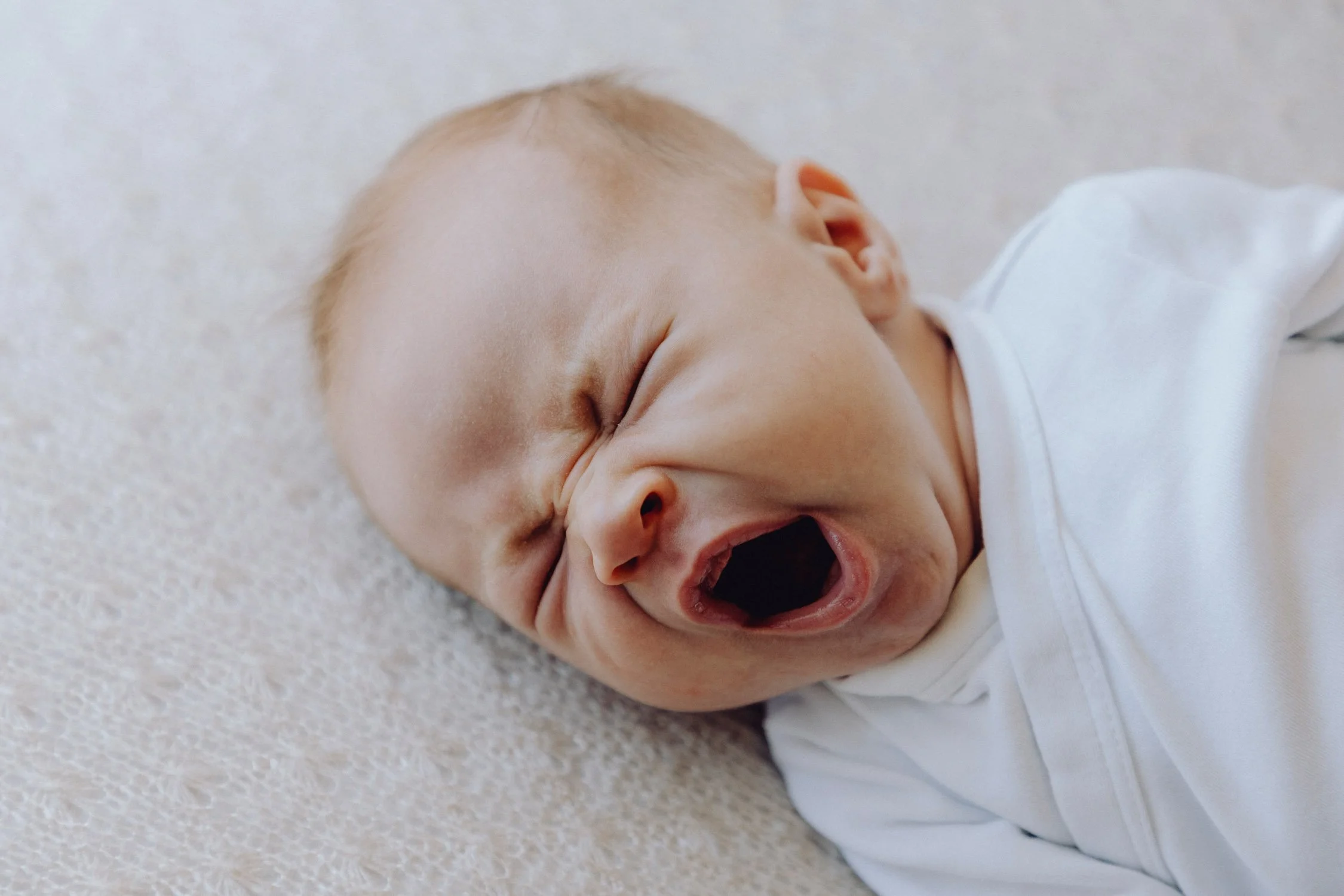 Ein schlafendes Baby liegt auf einer hellen, weichen Oberfläche und schreit mit offenem Mund.