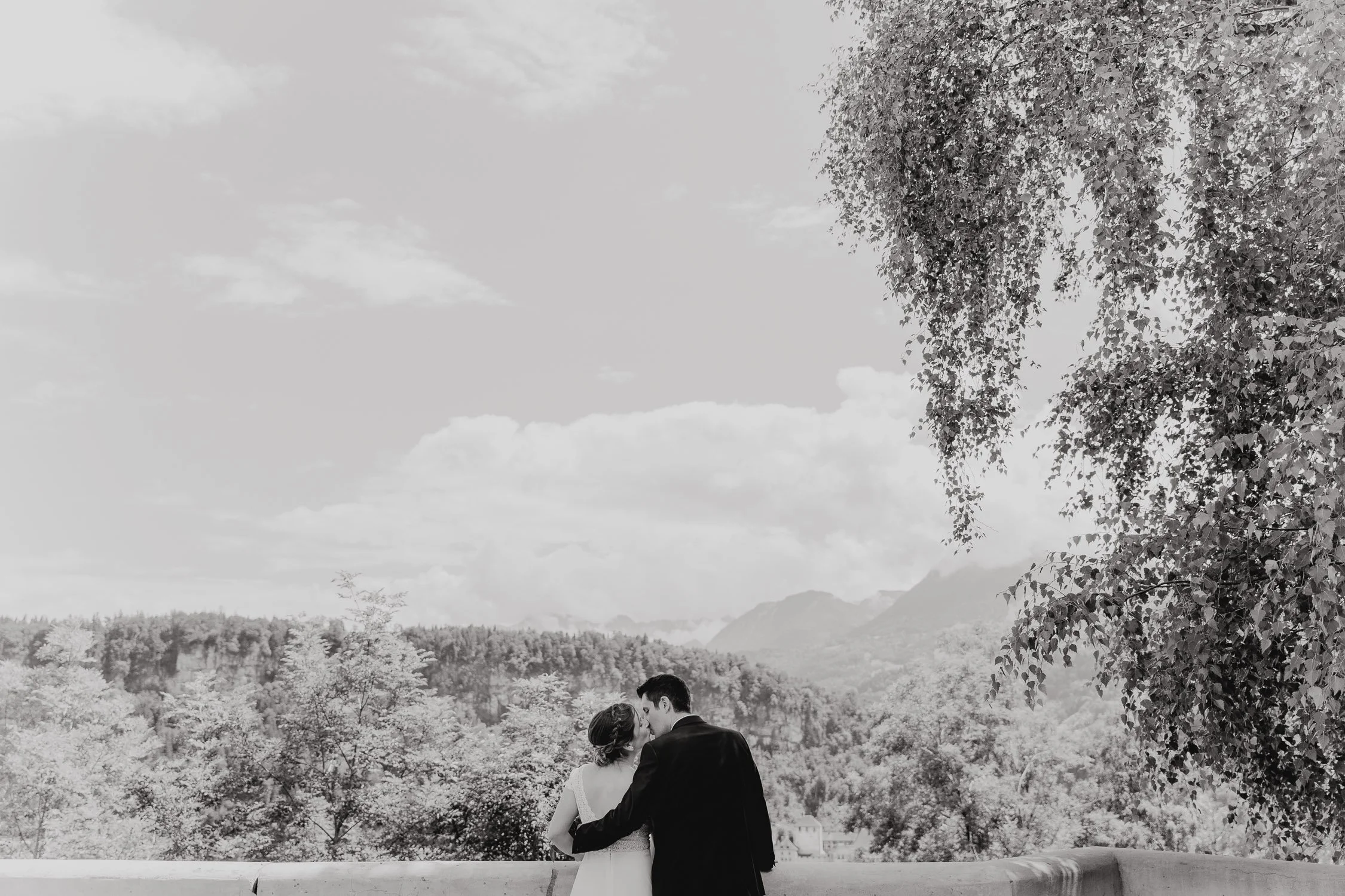 Ein Paar, wahrscheinlich Braut und Bräutigam, küsst sich vor einer Naturkulisse mit Bäumen und Bergen.
