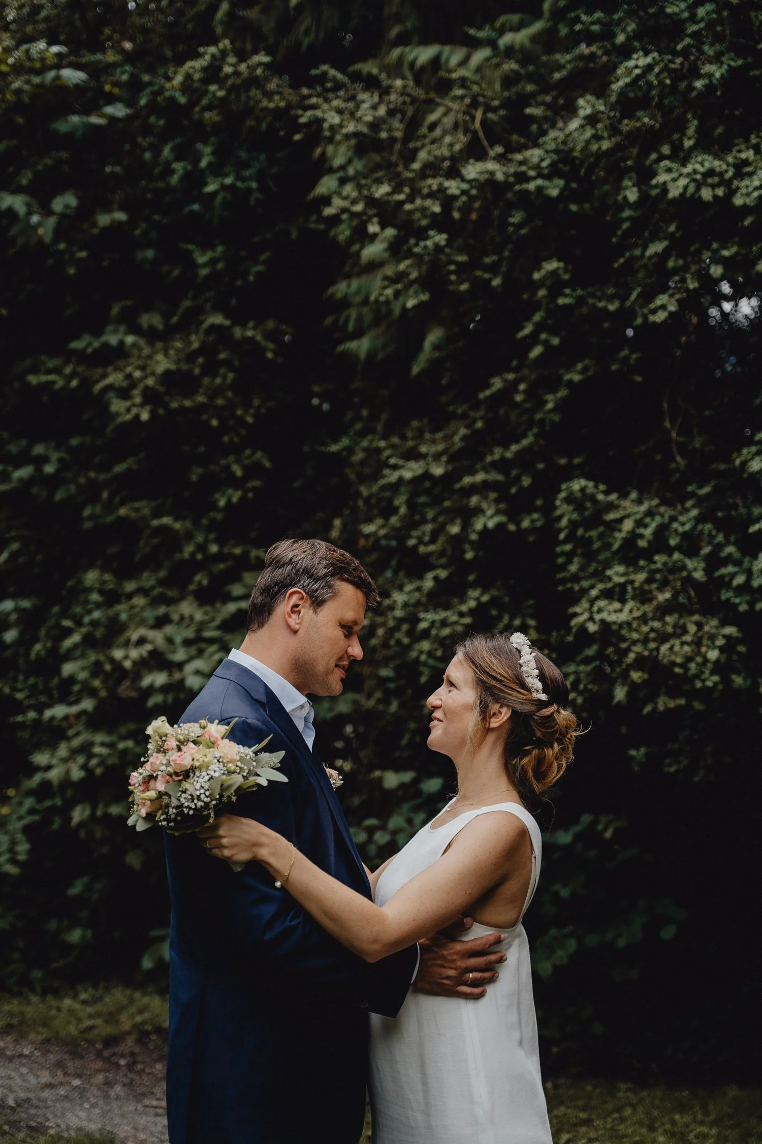Ein brautpaar steht sich gegenüber im Freien, umgeben von Bäumen, die sich liebevoll anschauen, sie hält einen Blumenstrauß, er trägt einen dunklen Anzug.