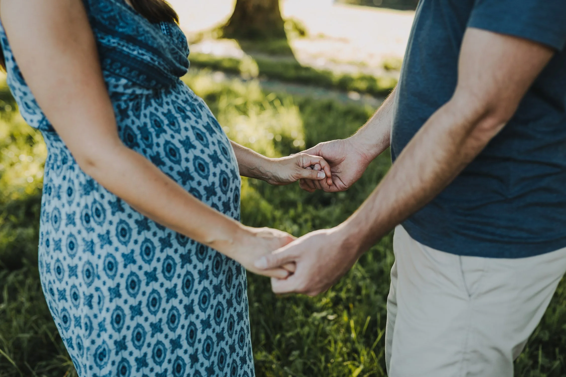 Ein Paar hält sich an den Händen und zeigt einen Schwangerschaftsbauch, draußen im Grünen, bei Sonnenlicht.
