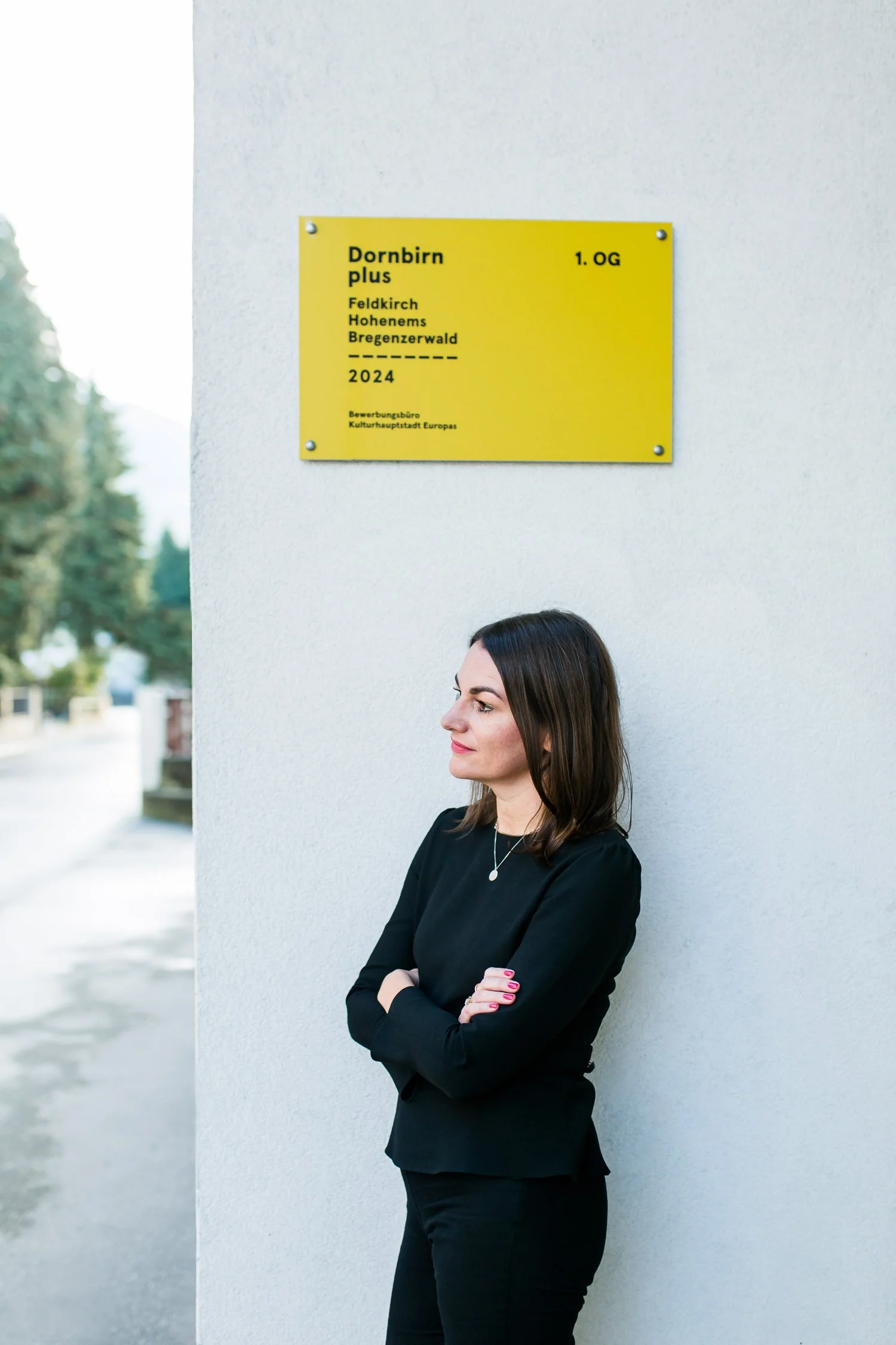 Eine Frau steht mit verschränkten Armen vor einer weißen Wand, über ihr eine gelbe Tafel mit schwarz gedrucktem Text. Der Text auf der Tafel zeigt Informationen zu einem Projekt namens 'Dornbirn plus' aus dem Jahr 2024.