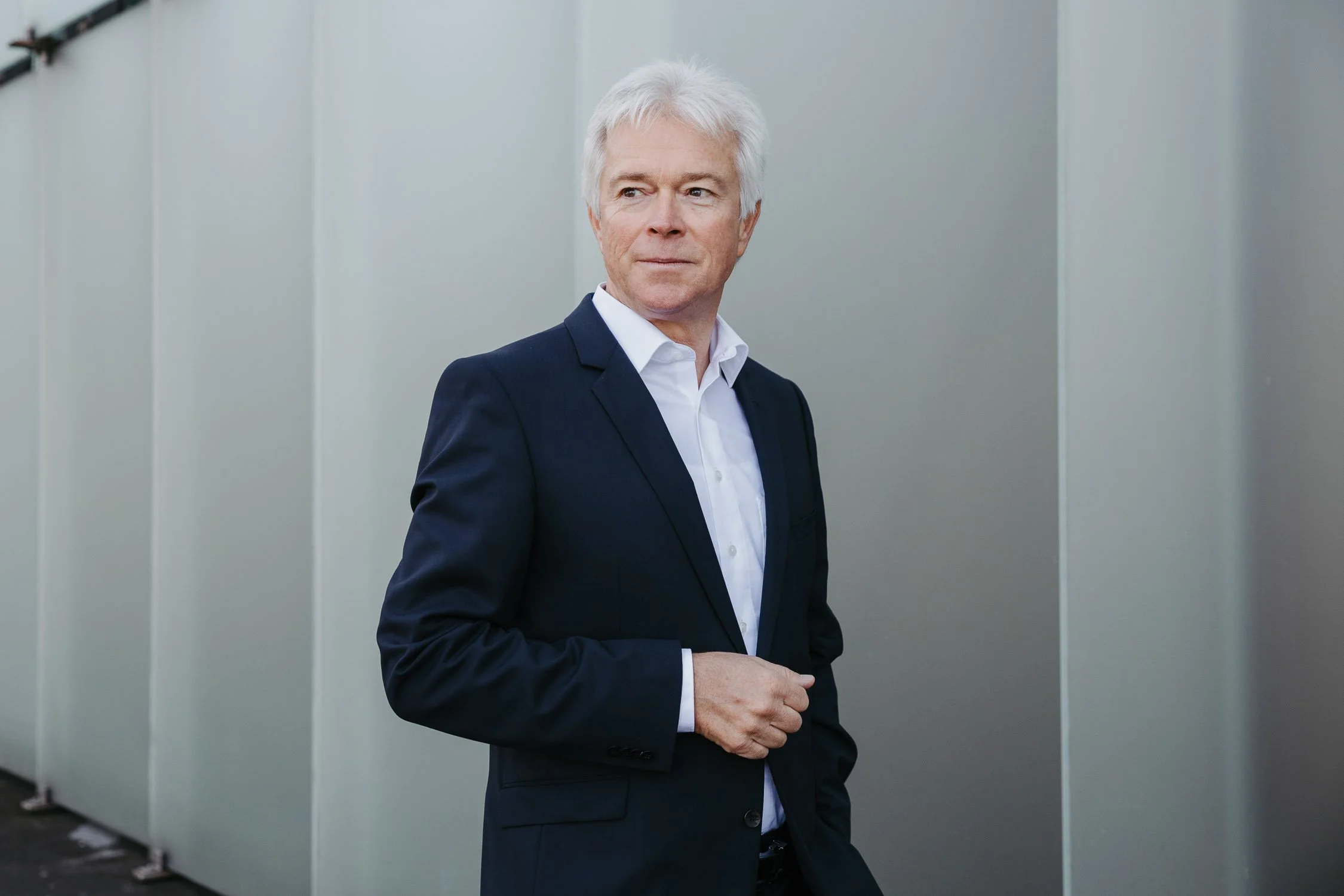 Ein älterer Mann in einem schwarzen Anzug steht vor einer grauen Wand und blickt zur Seite.