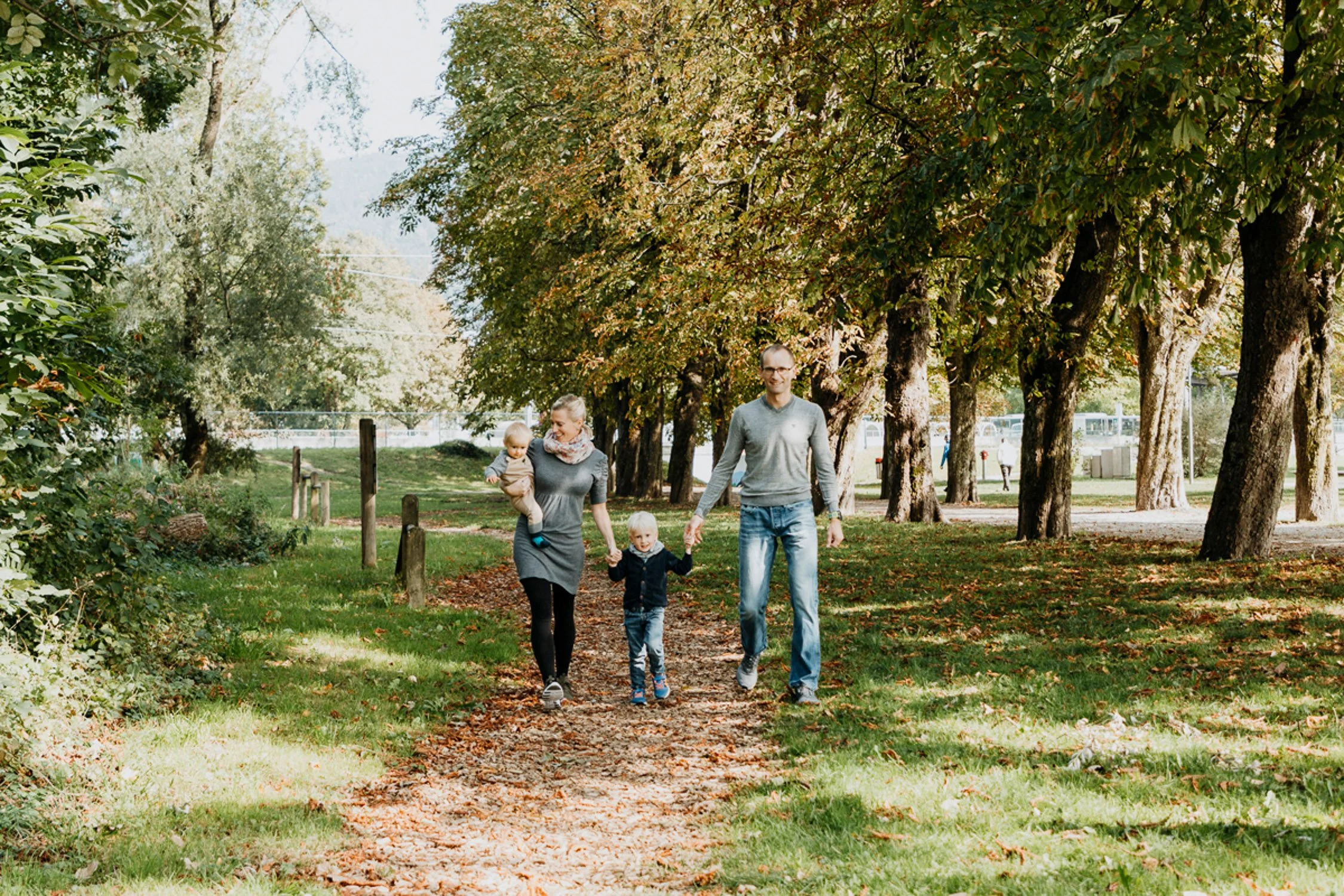 Familie, Vater, Mutter mit zwei kleinen Kindern, Spaziergang im Park an einem sonnigen Herbsttag, Bäume mit herbstlicher Laubfärbung, blauer Himmel.