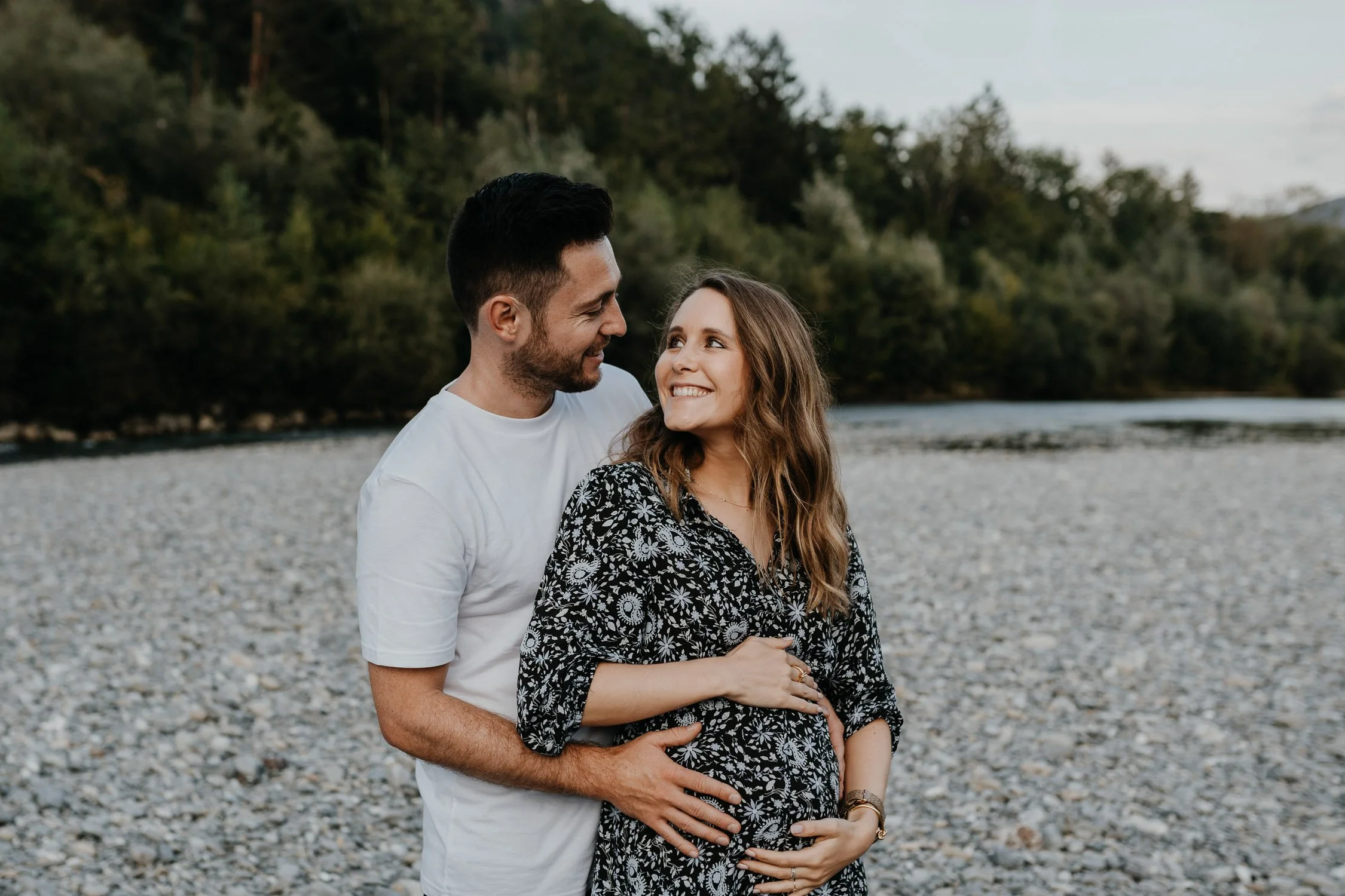 Ein Paar steht am Fluss, die schwangere Frau lächelt und der Mann umarmt sie liebevoll.