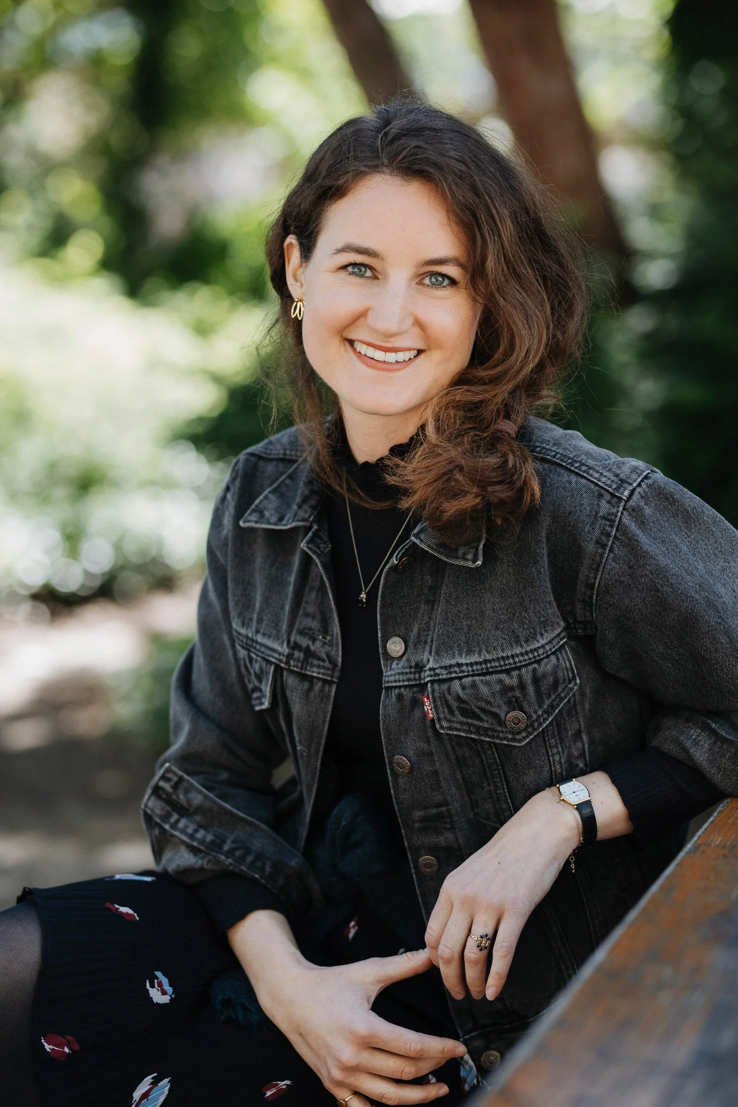 Eine lächelnde Frau mit braunen, lockigen Haaren sitzt auf einer Bank im Freien, umgeben von grünen Bäumen im Hintergrund.