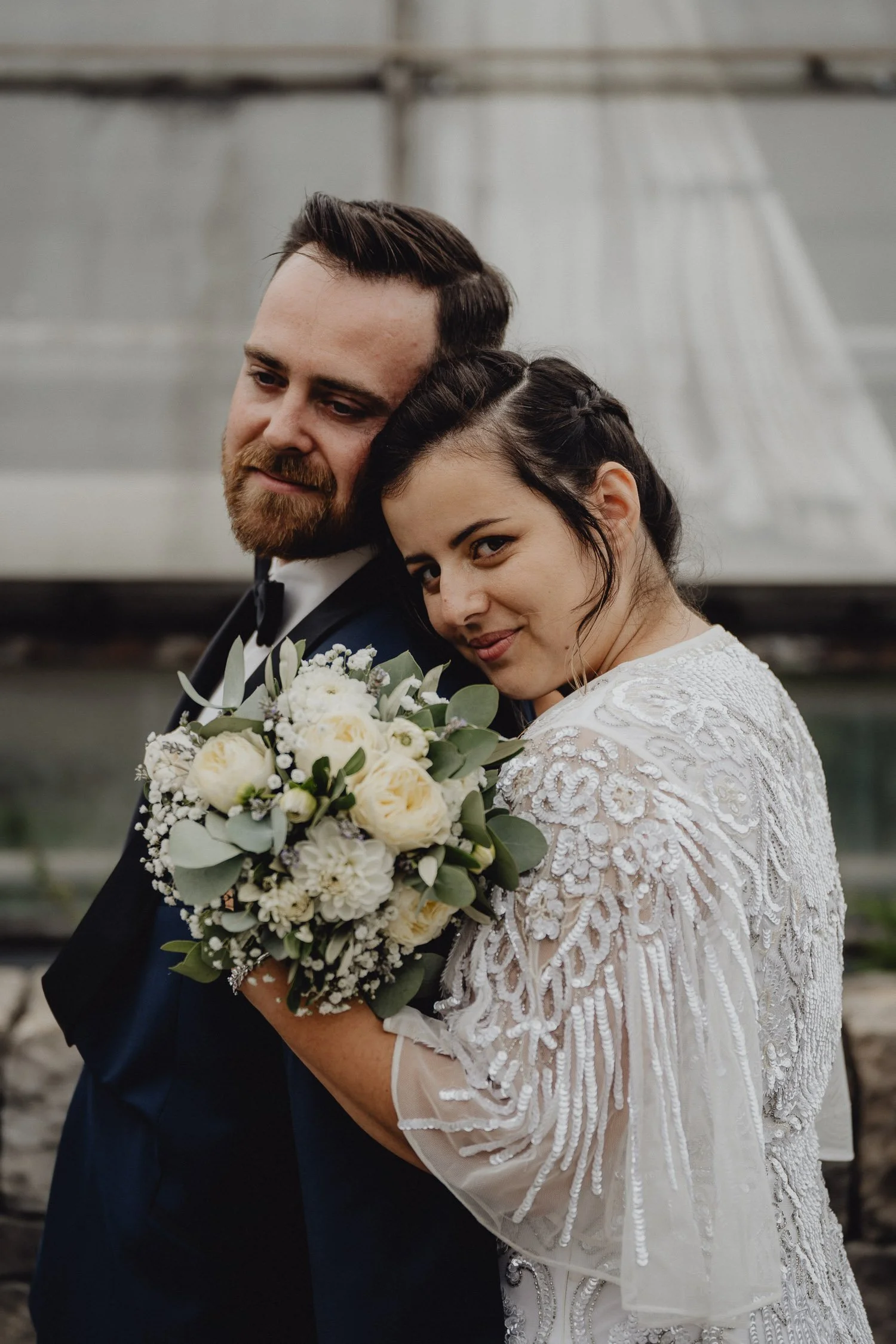 Ein Brautpaar posiert mit einem Blumenstrauß während einer Hochzeit, draußen vor einer Glasscheibe.