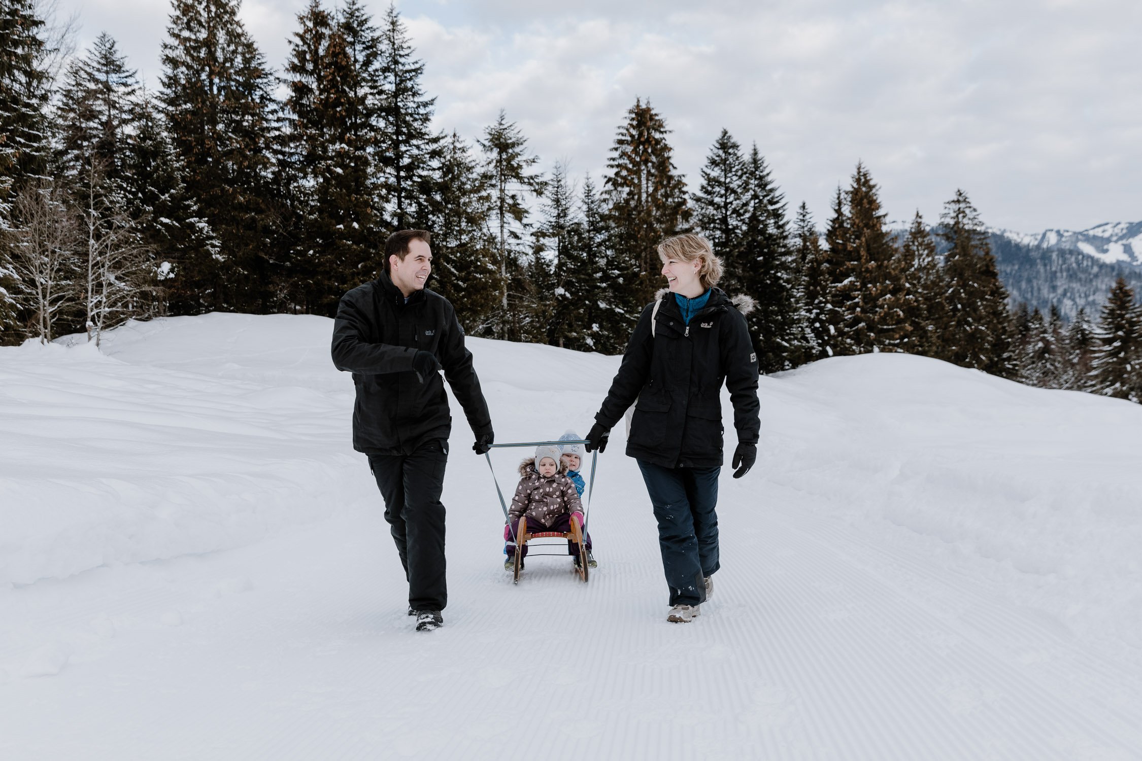 Zwei Erwachsene ziehen mit einer roten Schlitten zwei kleine Kinder durch den Schnee, umgeben von Tannenbäumen und Berge, bei winterlichem Himmel.