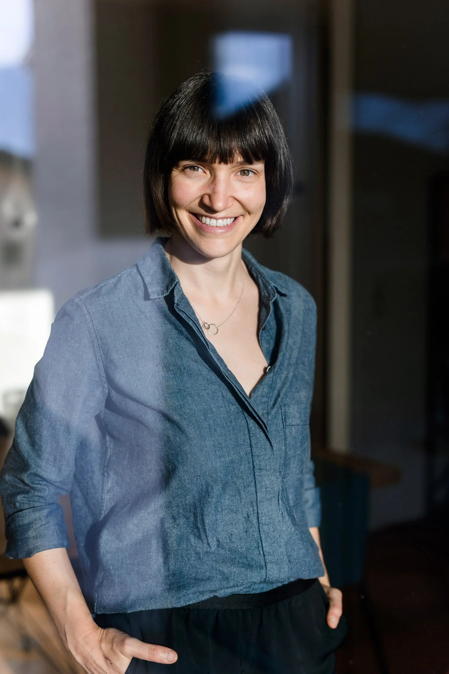 Eine lachende Frau mit dunklem, kurzem Haarschnitt trägt eine blaue Jeansjacke und schwarze Hosen, steht vor einem dunklen Hintergrund. Hochwertiges Porträt, natürliches Licht.