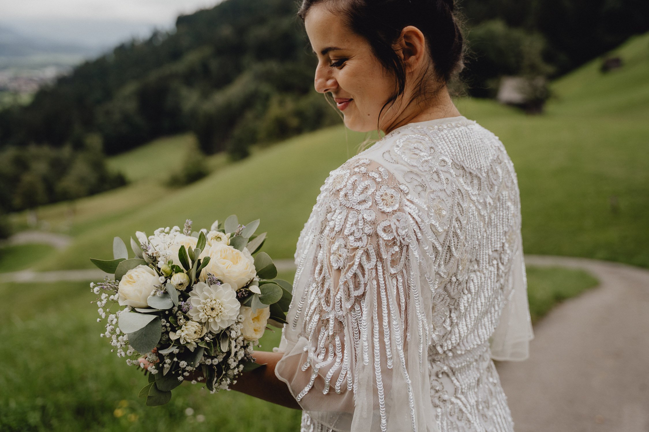 Eine Frau im weißen Hochzeitskleid hält einen Blumenstrauß aus weißen Rosen, Blüten und grünem Laub, während sie in einer grünen Hügellandschaft steht.