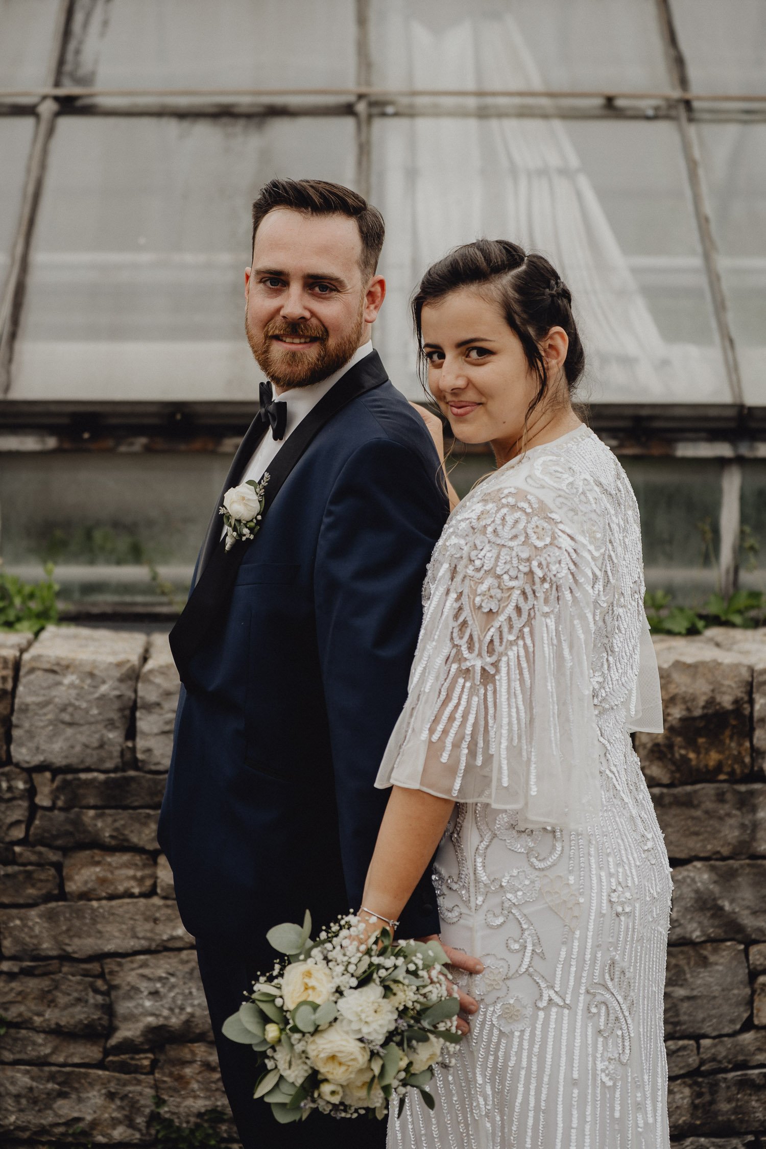 Ein Brautpaar steht nebeneinander vor einem Steinmauer und einer Glashausterrasse, der Bräutigam trägt einen schwarzen Anzug mit Fliege, die Braut trägt ein weißes, handbesticktes Brautkleid und hält einen Hochzeitsstrauß aus weißen Blumen und grünem