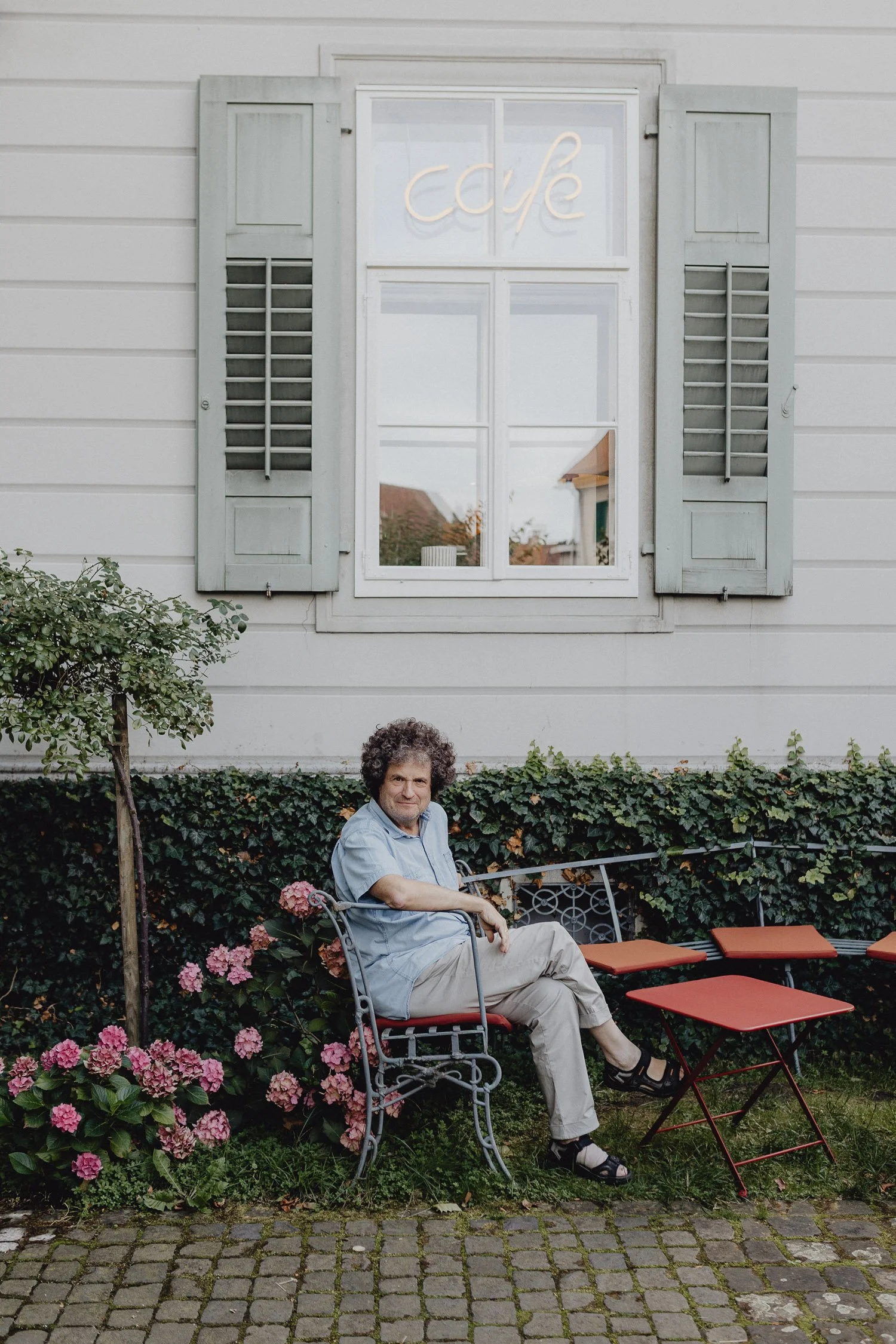 Ein älterer Mann sitzt auf einer Gartenbank vor einem weißen Haus mit Fenster und blauen Fensterläden, die terrakottafarbene Sitzkissen auf bequemen Stühlen, umgeben von Blumen und grünen Pflanzen.
