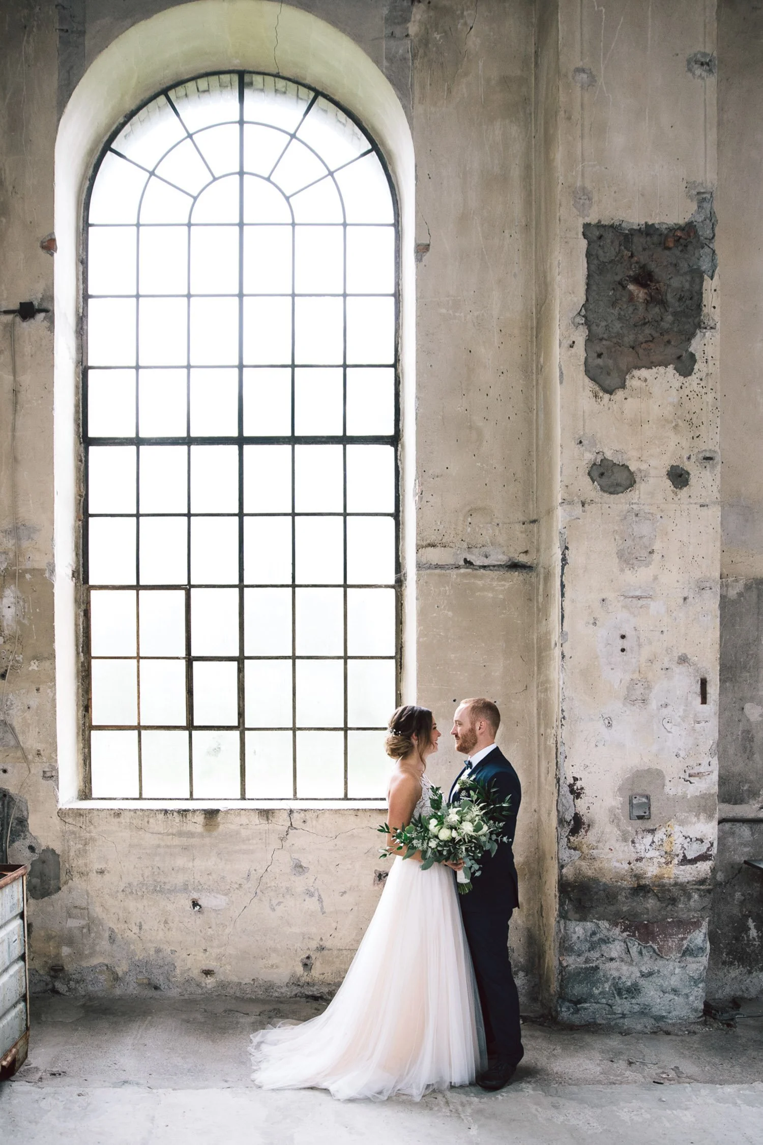 Ein Brautpaar steht sich in einer alten, verfallenen Fabrikhalle gegenüber. Die Braut trägt ein langes, weißes Hochzeitskleid und hält einen großen Blumenstrauß, während der Bräutigam einen dunklen Anzug trägt. Im Hintergrund ein großes, altes Fenste