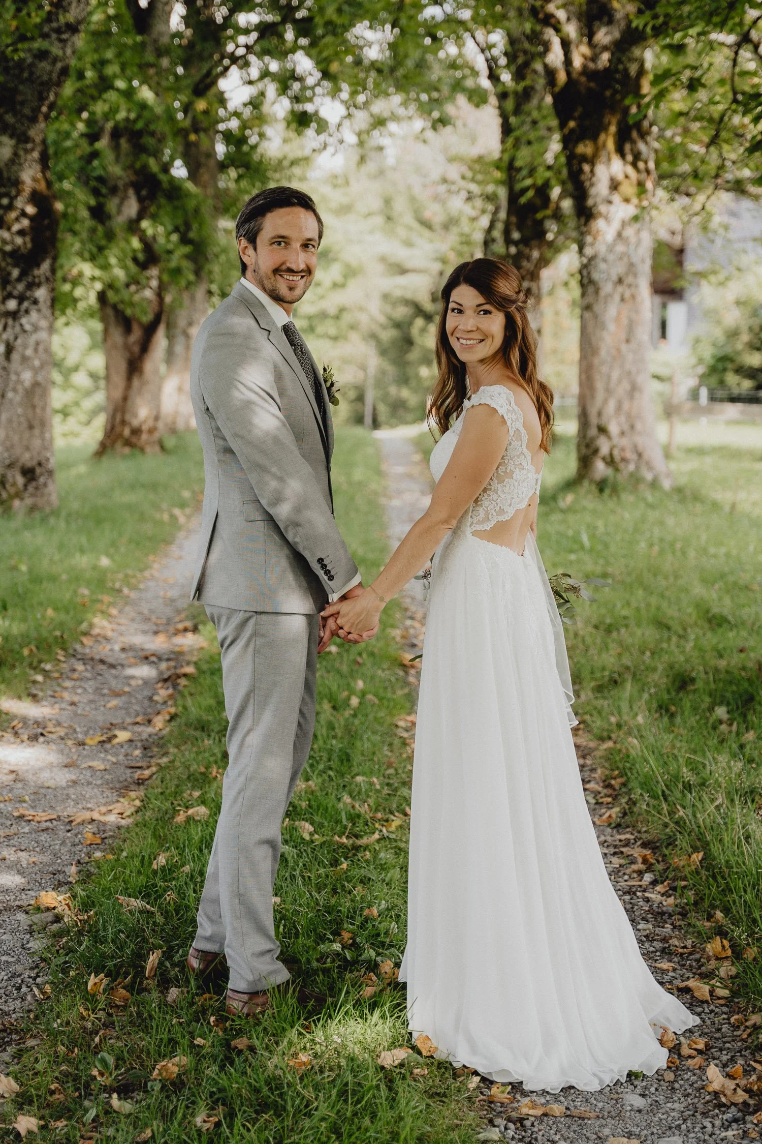 Ein brautpaar hält sich an einer Baumallee an den Händen, vermissen ein Lächeln. Sie tragen formelle Kleidung, sie in einem weißen Brautkleid und er in einem grauen Anzug. Die Natur um sie herum ist grün und sonnig.