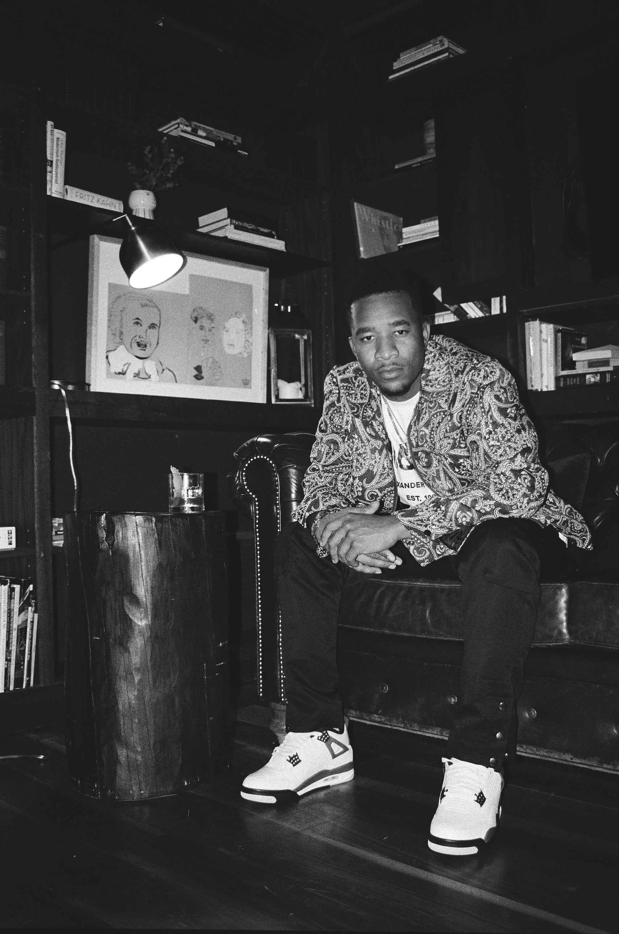 A young man sitting on a leather couch in a dimly lit room with bookshelves, a framed cartoon picture, and a table, wearing a patterned jacket, white sneakers, and posing with a serious expression.