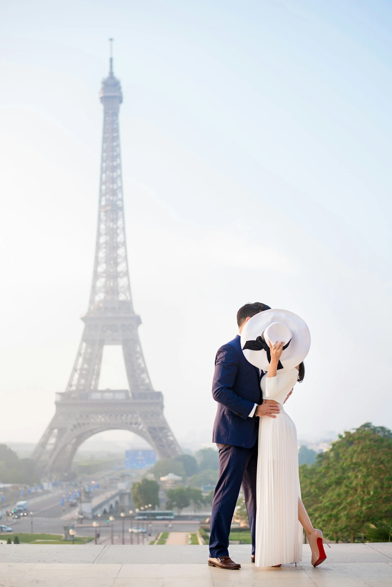 A couple embracing in front of the Eiffel Tower in Paris, with the woman holding a large sun hat and wearing red high heels.