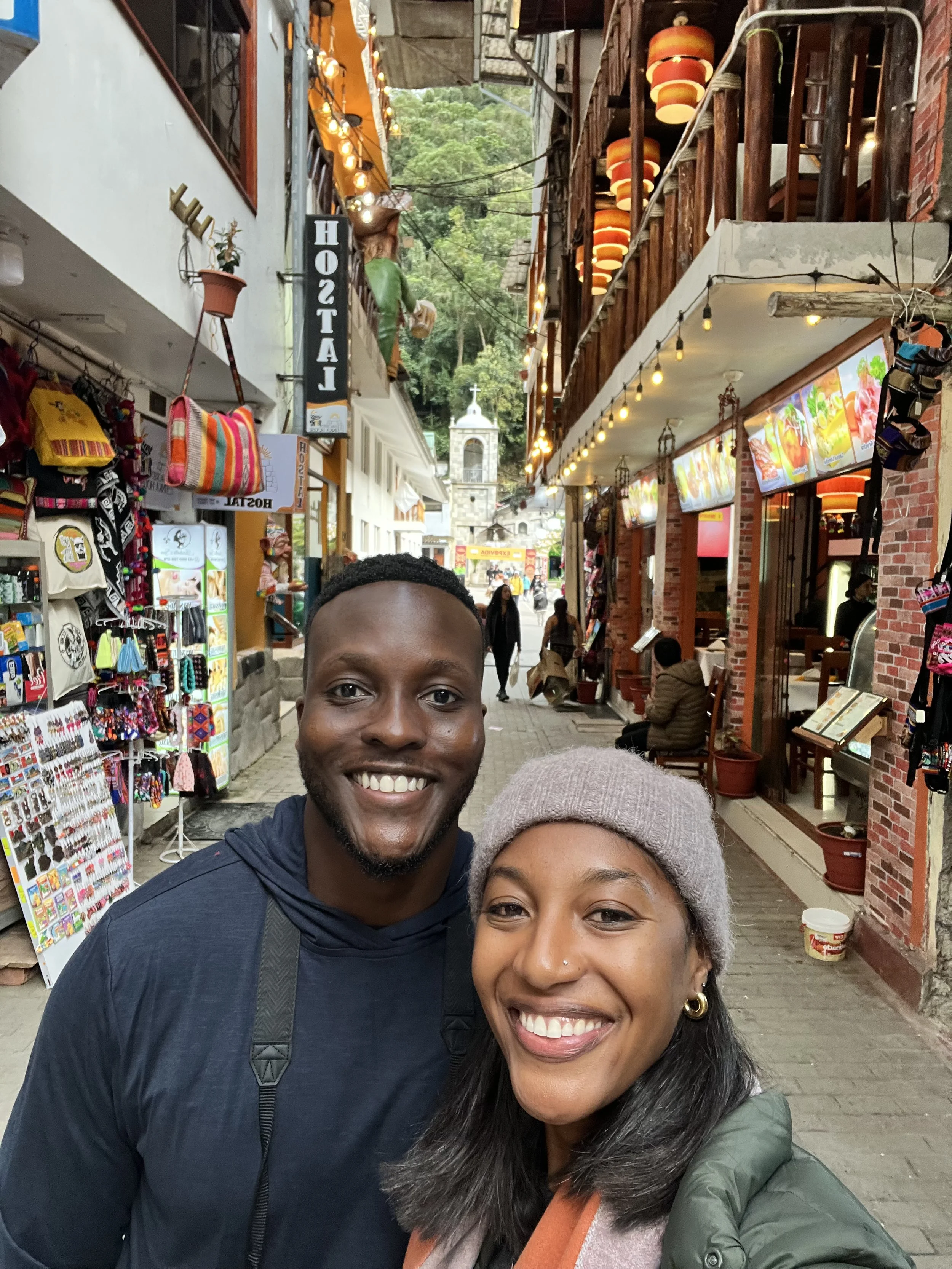 A selfie of a smiling man and woman standing close together on a busy street with shops and hanging lanterns, with scenic greenery and a church in the background.