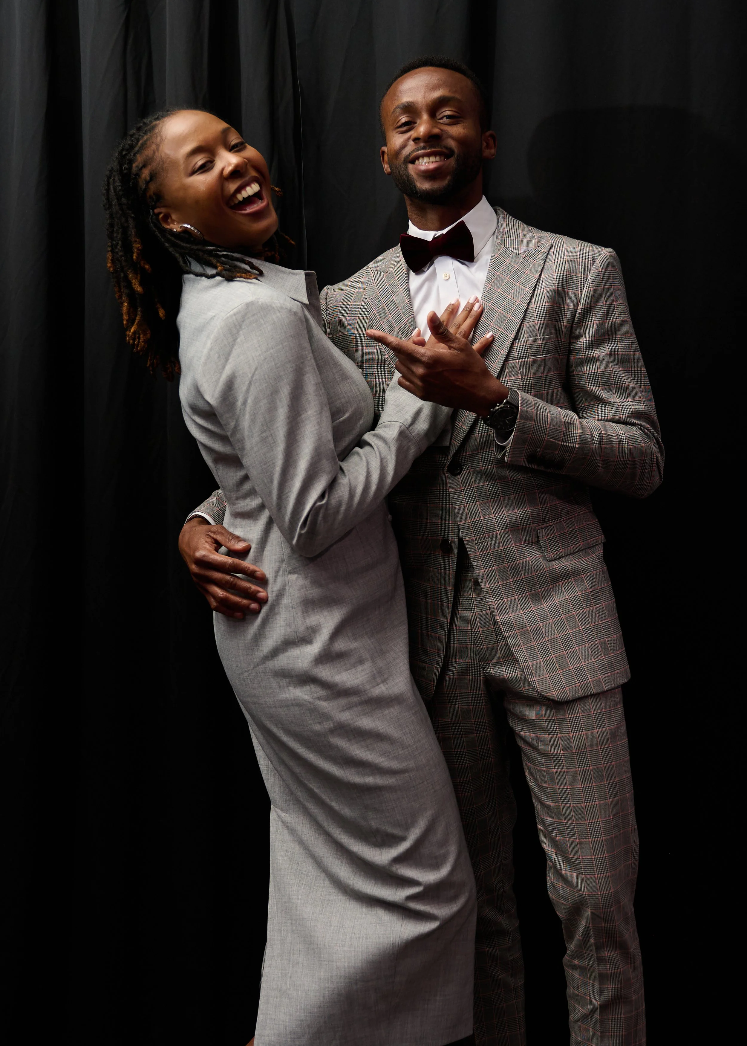 A smiling woman and a smiling man dressed in formal attire, standing against a black background. The woman has dreadlocks and is wearing a light gray suit, while the man is wearing a plaid suit with a bow tie and watch.