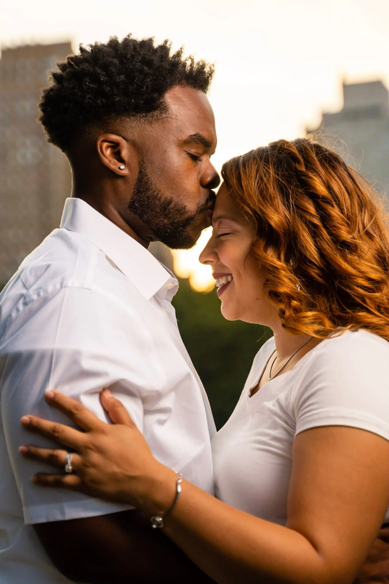 A couple is embracing outdoors during sunset, with their foreheads touching and eyes closed, smiling.