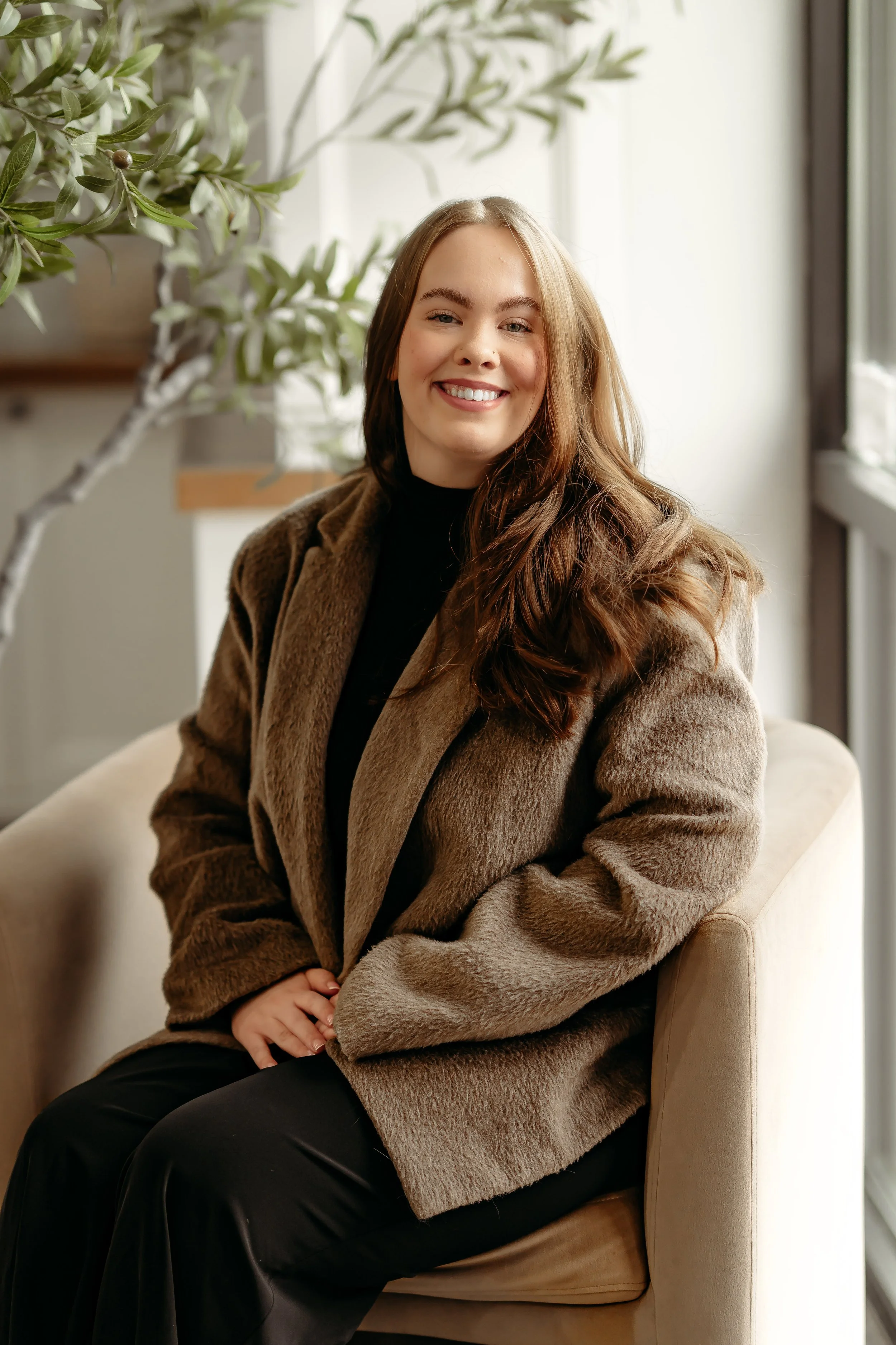 A woman with long brown hair sitting on a beige chair next to a window, wearing a brown fur coat and black pants, smiling indoors with green plants in the background.