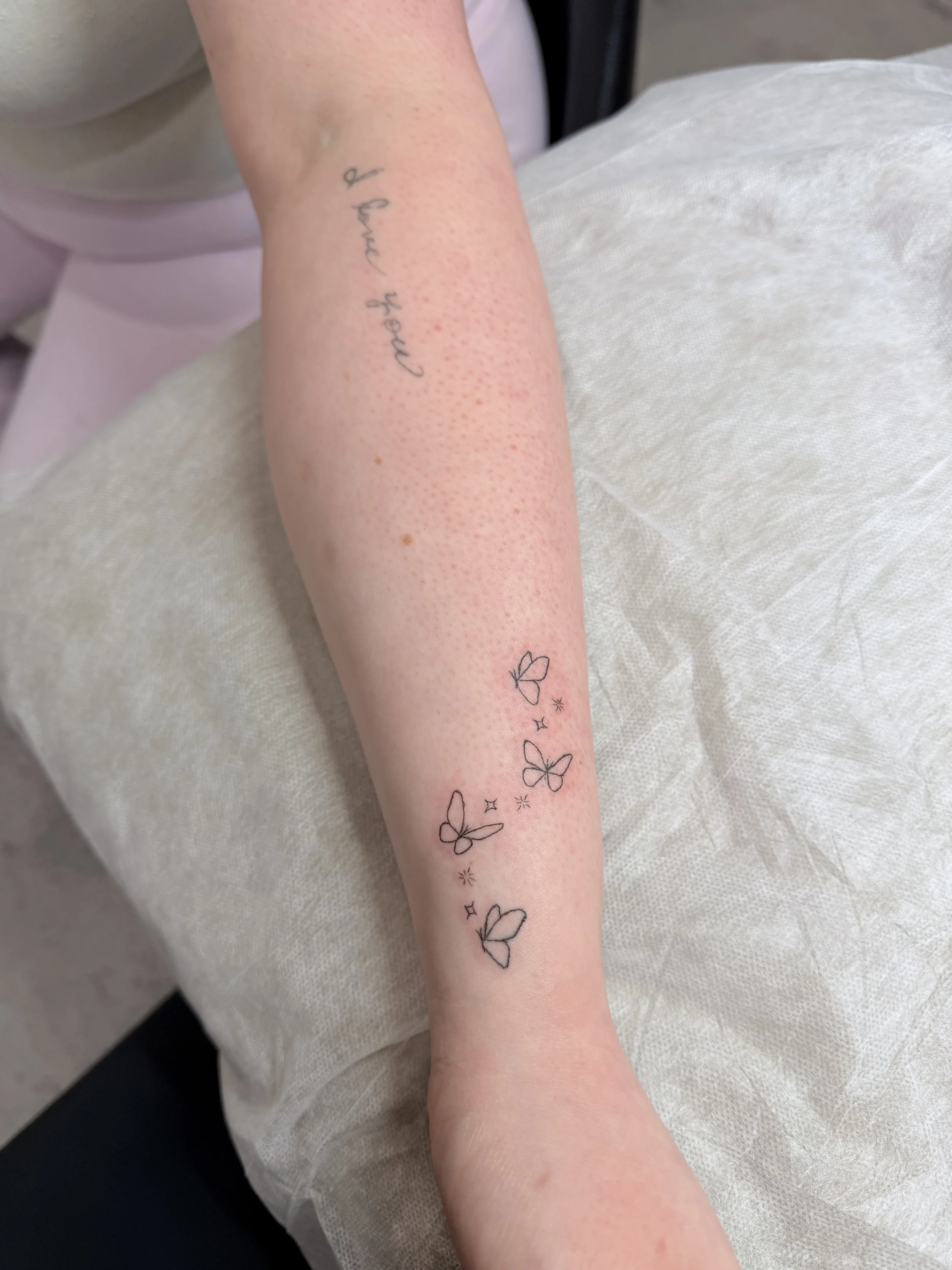 A person's forearm with a tattoo of butterflies and stars, and the faint text 'of pure gold'.