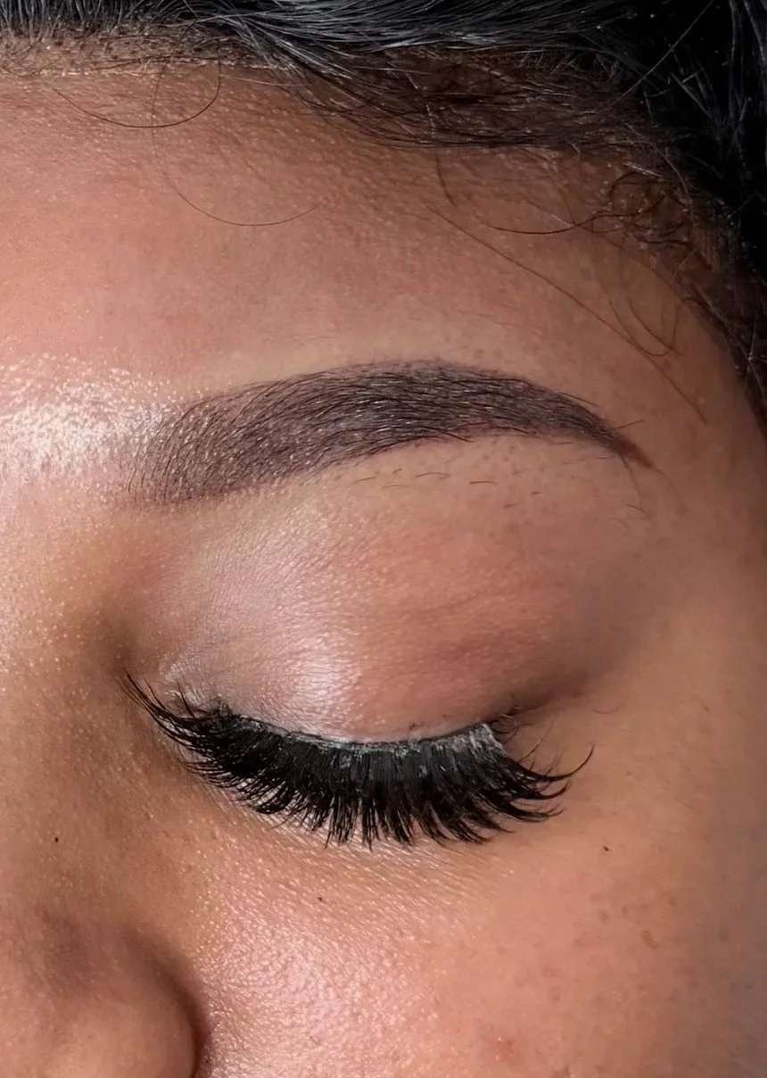 Close-up of a woman's closed eye showing well-groomed eyebrows, long curled eyelashes, and smooth skin with natural makeup.