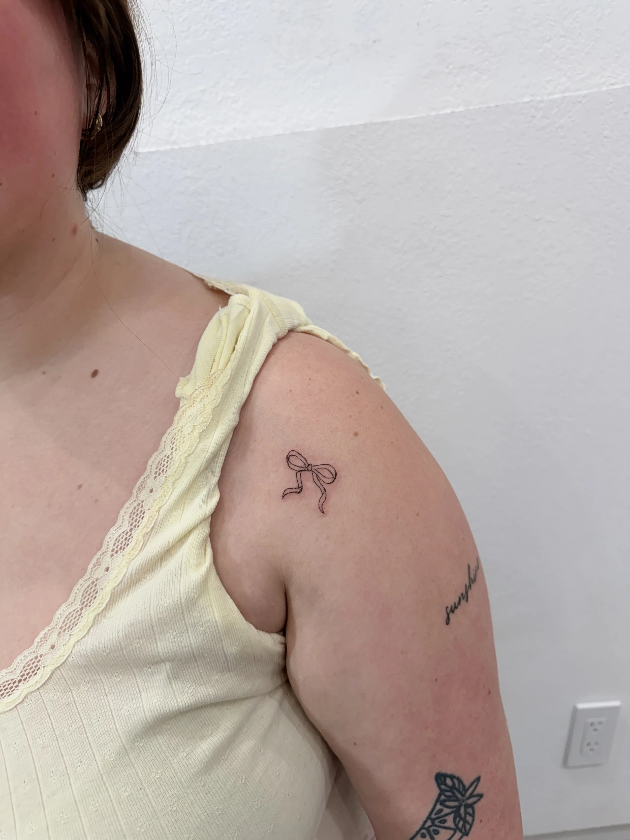 A close-up of a woman's shoulder and upper arm, showing a tattoo of a bow on her shoulder and a tattoo of a leaf on her forearm. She is wearing a light yellow top with lace details.