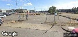 Empty parking lot with chains blocking access, situated near a city street with buildings in the background.