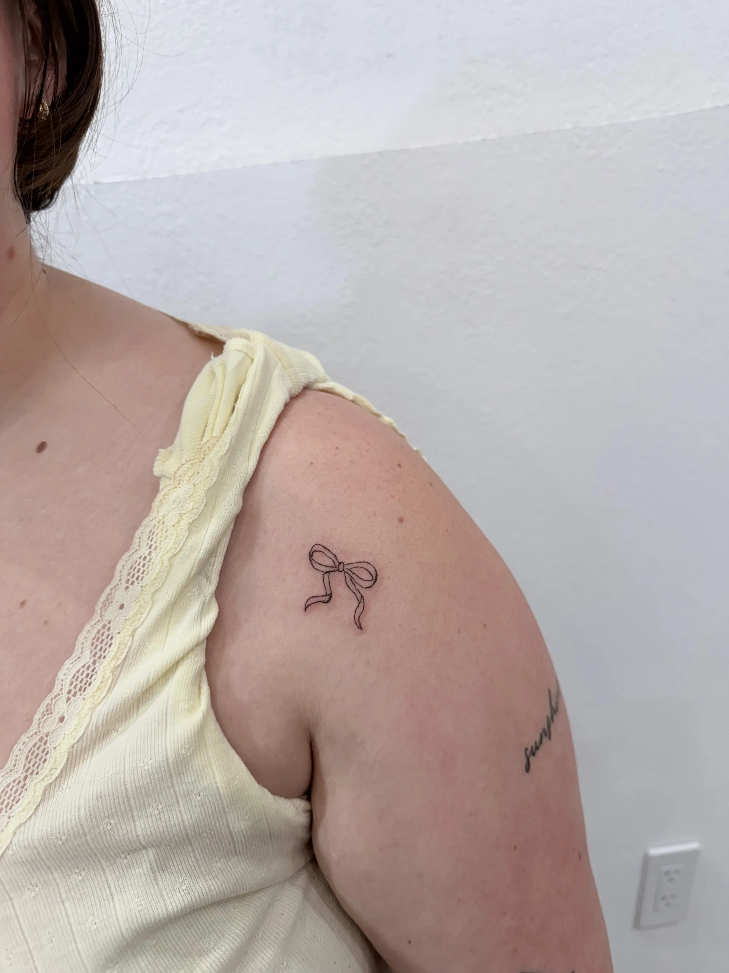 Close-up of a woman's upper arm and shoulder showing a black tattoo of a bow, with part of her face and ear visible. She wears a yellow tank top with lace trim.