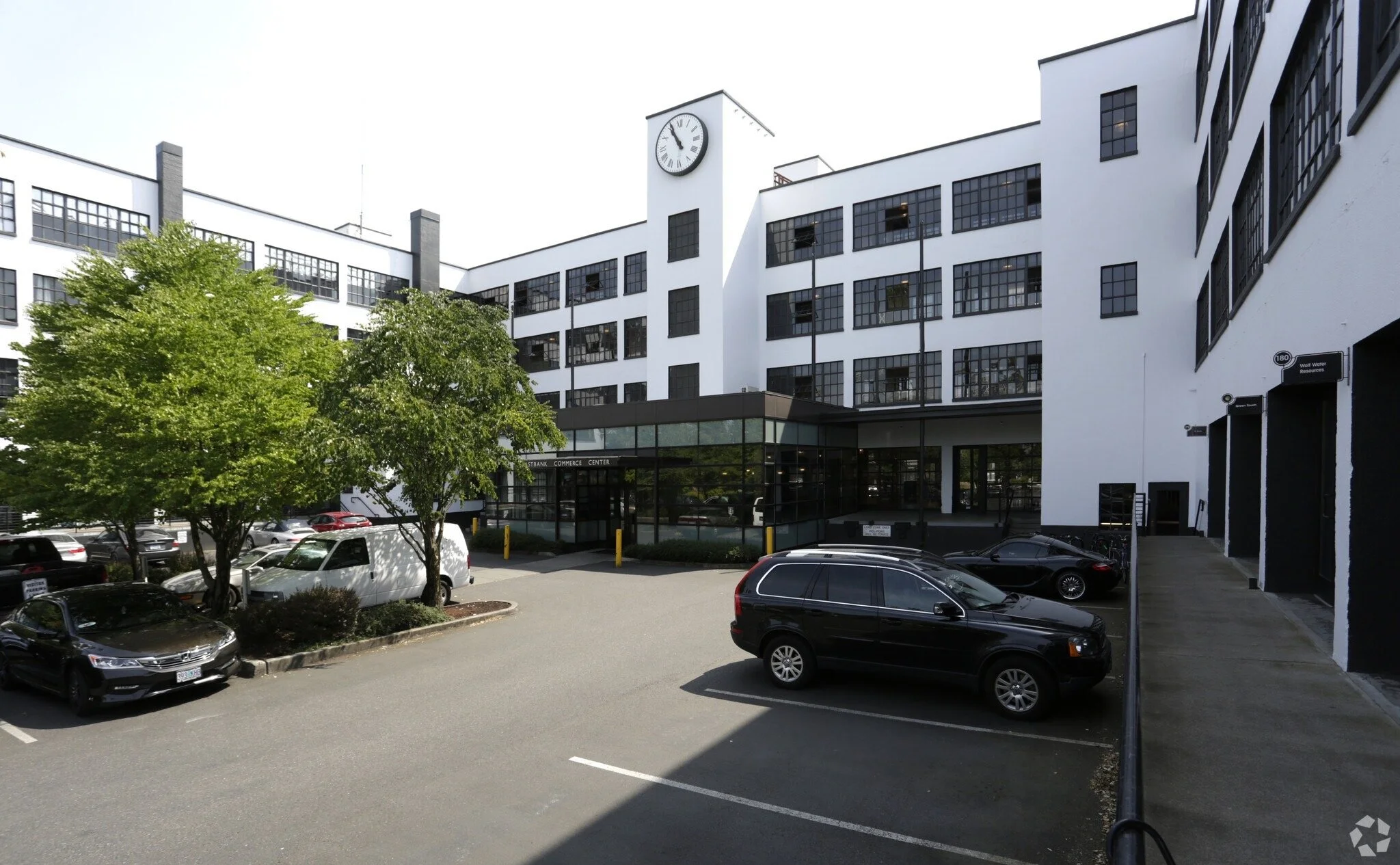 A parking lot in front of a modern white building with numerous black-framed windows and a clock tower showing approximately 9:05. Several cars are parked, and there are green trees and street signs.