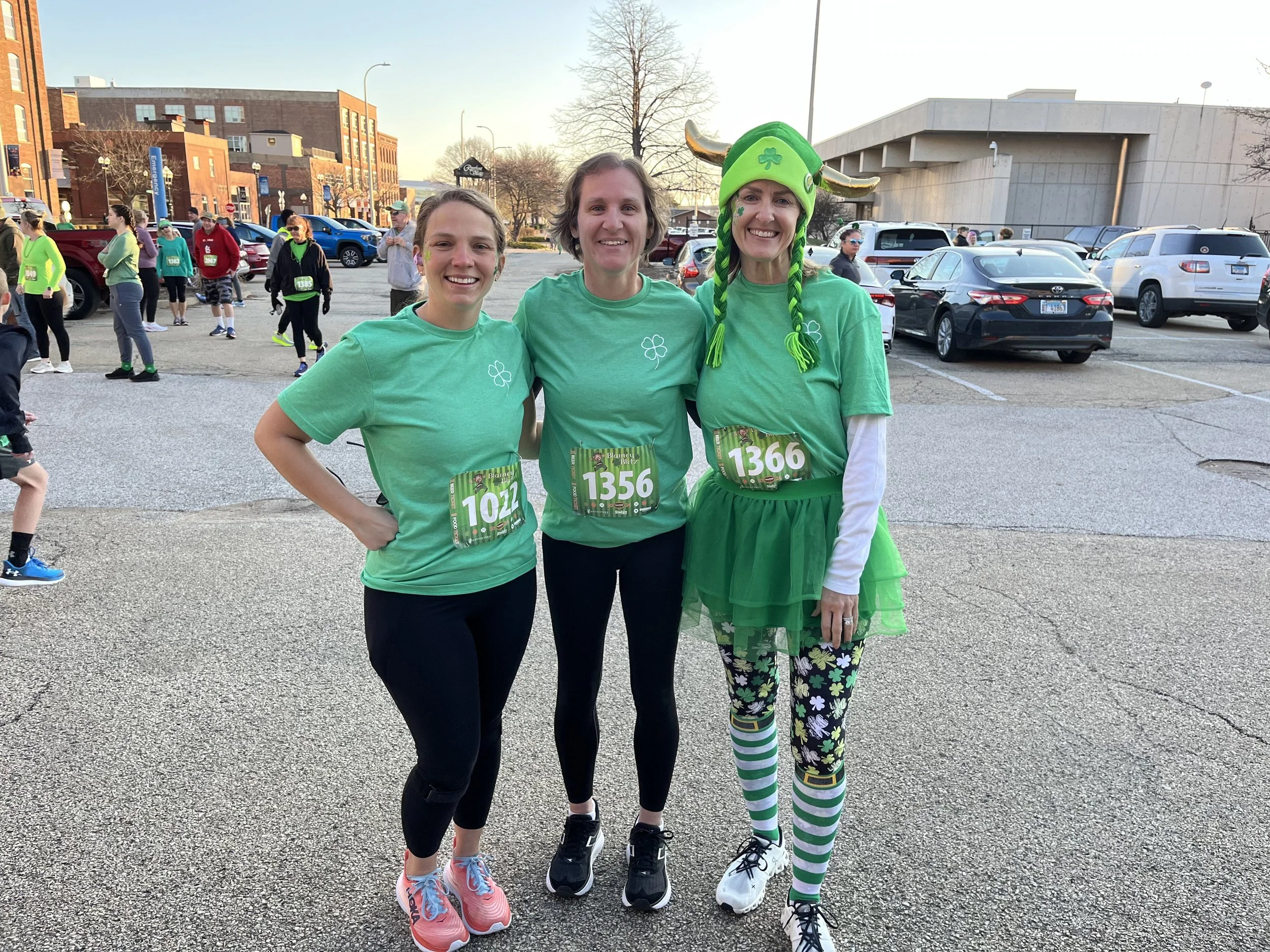 Three women with race bibs and runner attire standing in a parking lot during a St. Patrick's Day event, one dressed as a leprechaun.