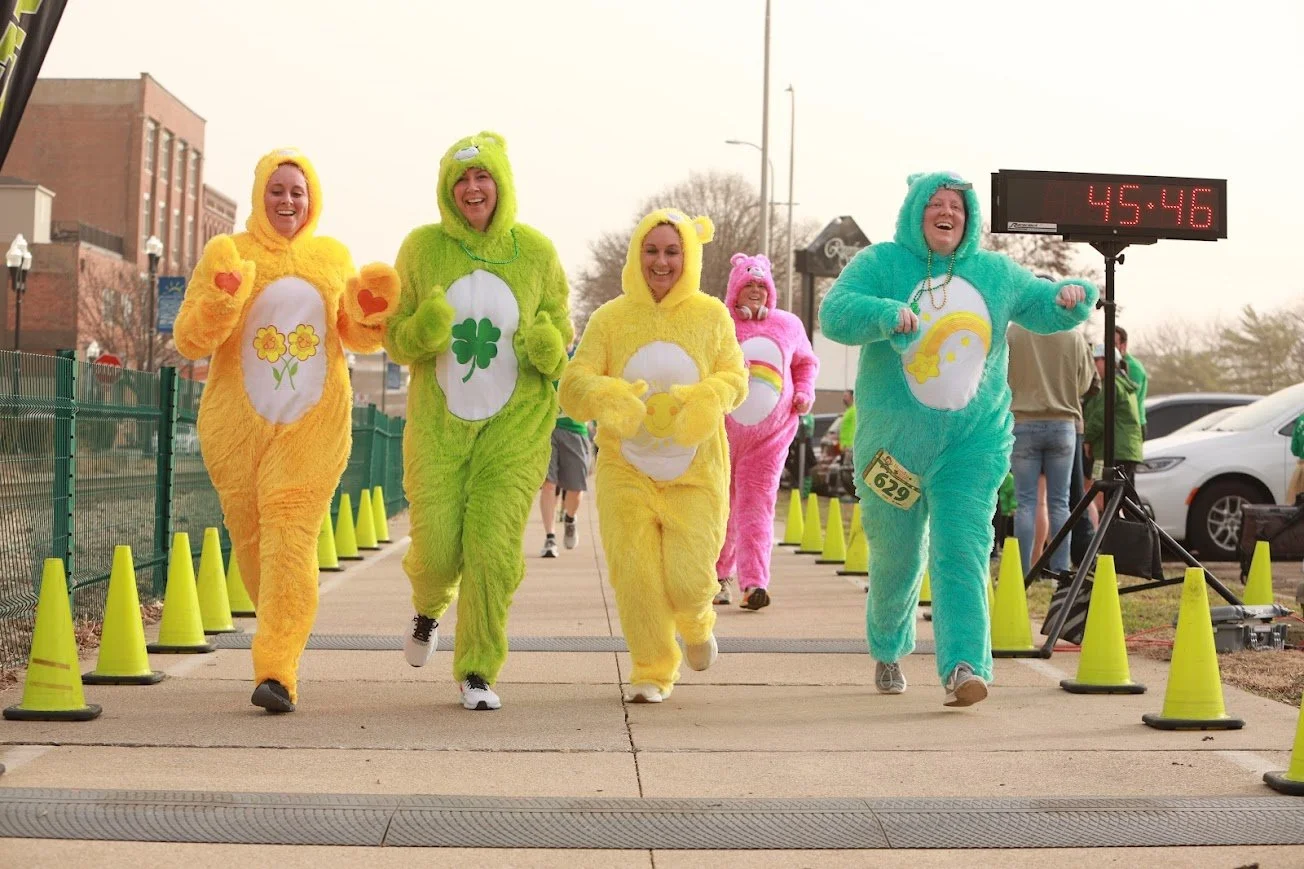 Group of people dressed in colorful plush animal costumes, running in a race event with a digital timer showing 45:46 on a bright day.