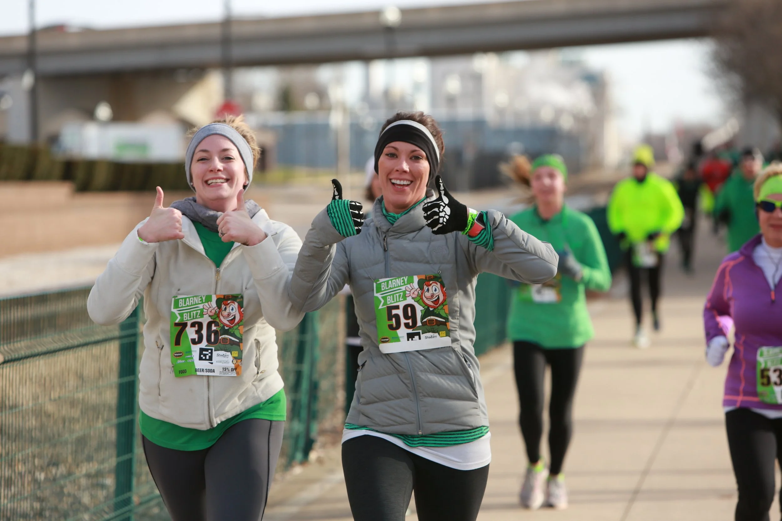 Group of runners participating in a race outdoors, wearing colorful athletic clothing and race bibs, smiling and giving thumbs-up signs.