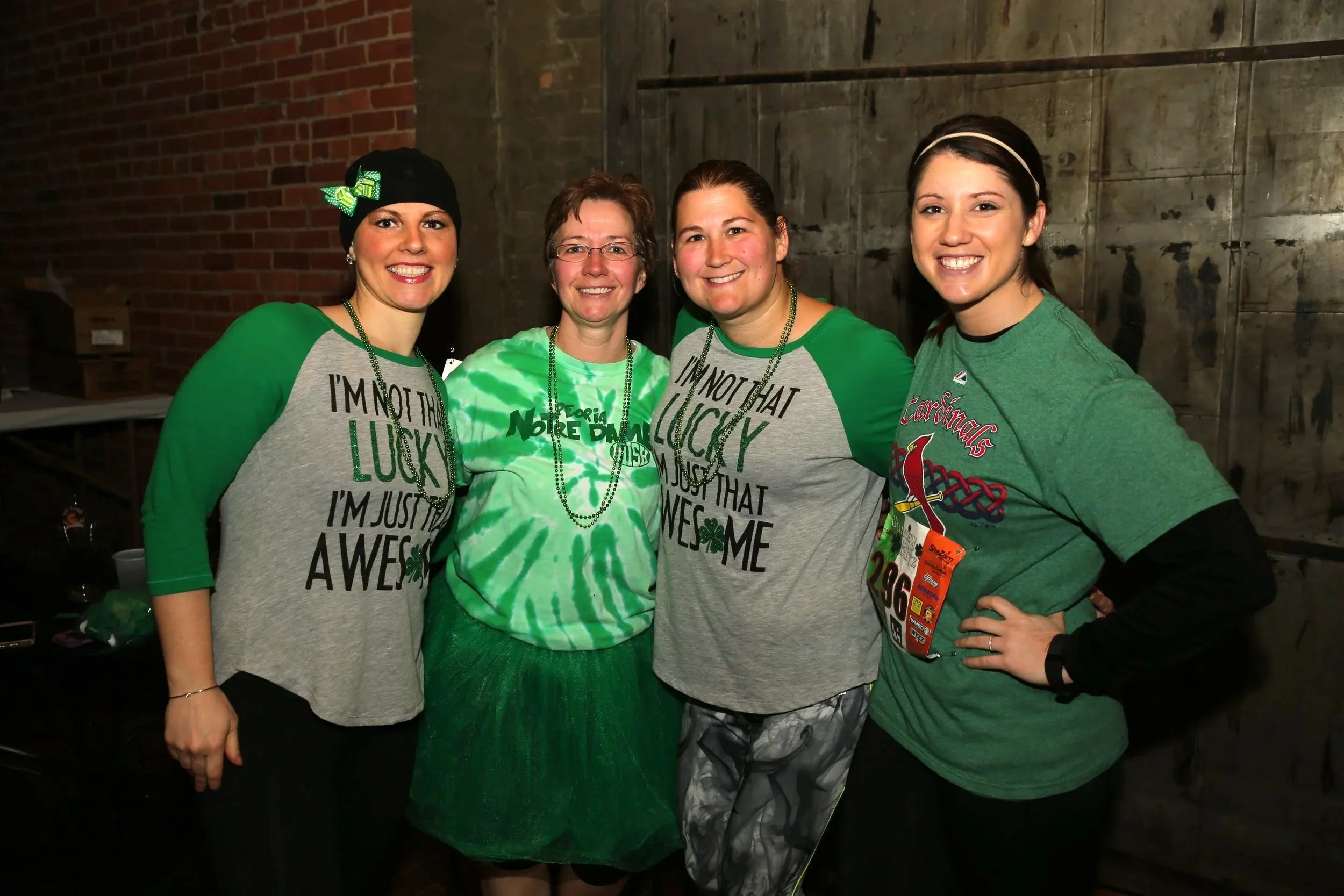Four women standing together indoors, dressed in green and gray shirts celebrating St. Patrick's Day, smiling at the camera.
