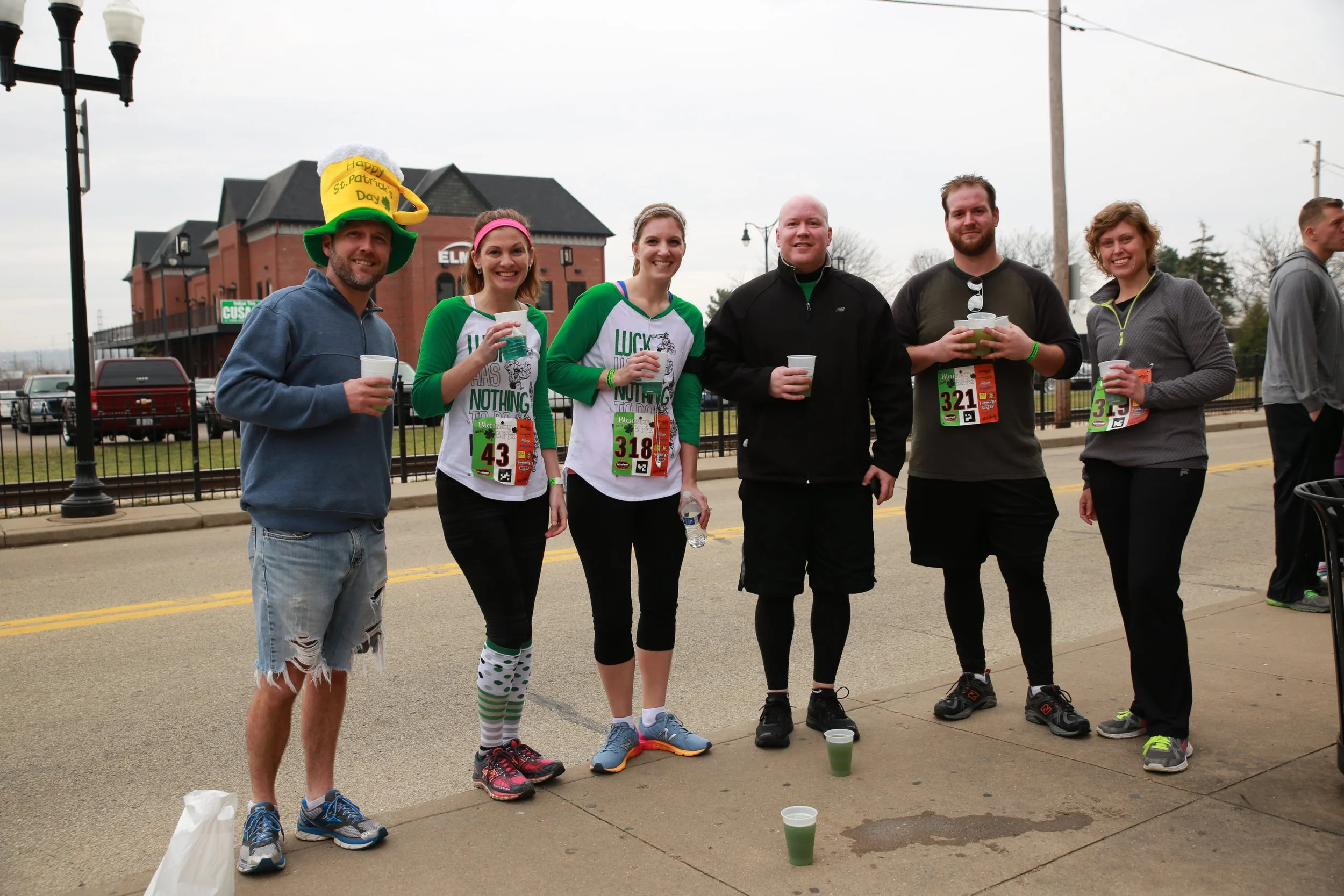 A group of six people standing on a sidewalk during a race, holding cups, some with race bibs, and dressed in athletic clothing. One person is wearing a large, colorful hat with a message for St. Patrick's Day.
