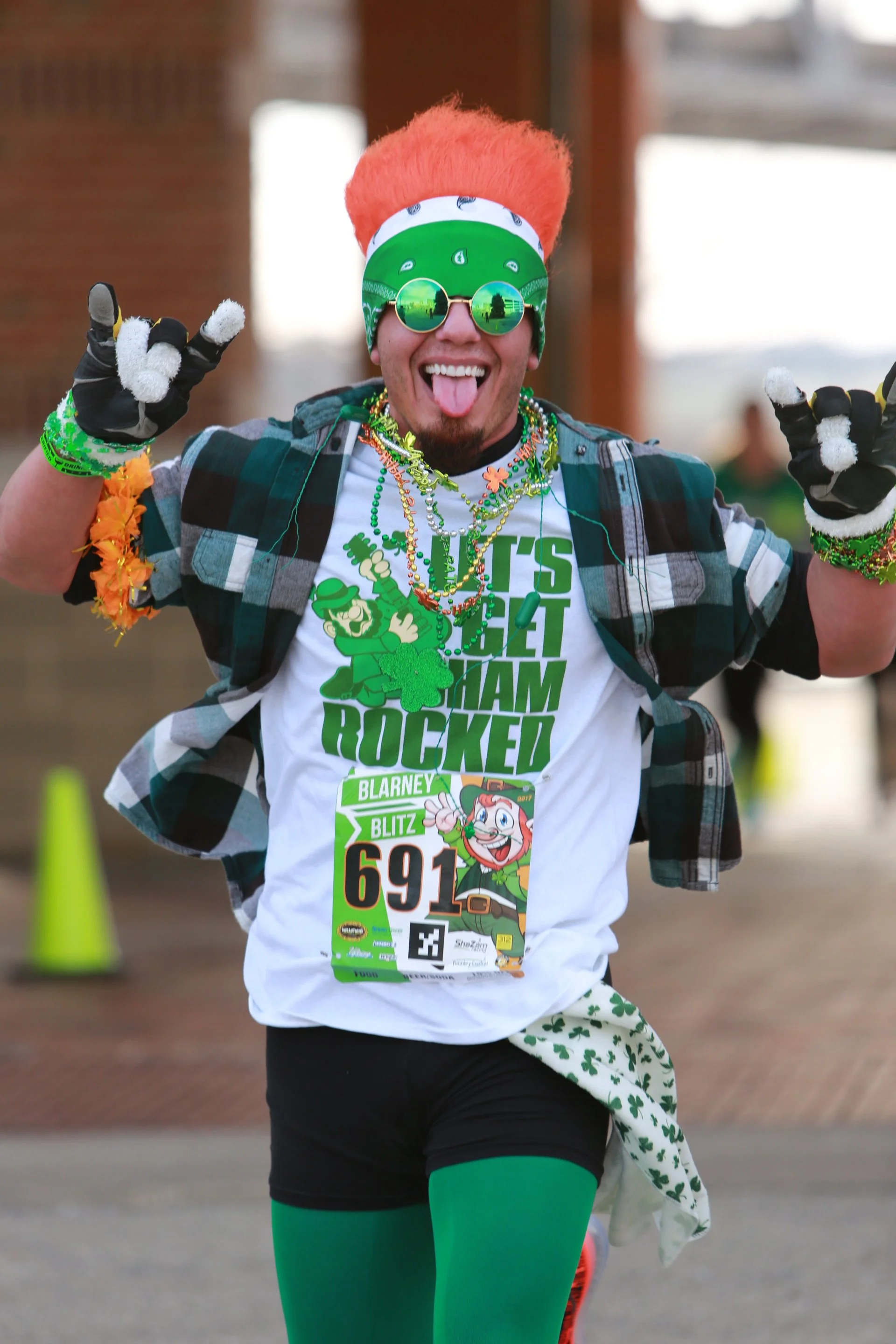 A man with bright orange hair, wearing green-tinted glasses, a green headband, and festive Mardi Gras accessories, joyfully celebrating with a wide smile and tongue sticking out during a St. Patrick's Day themed event.