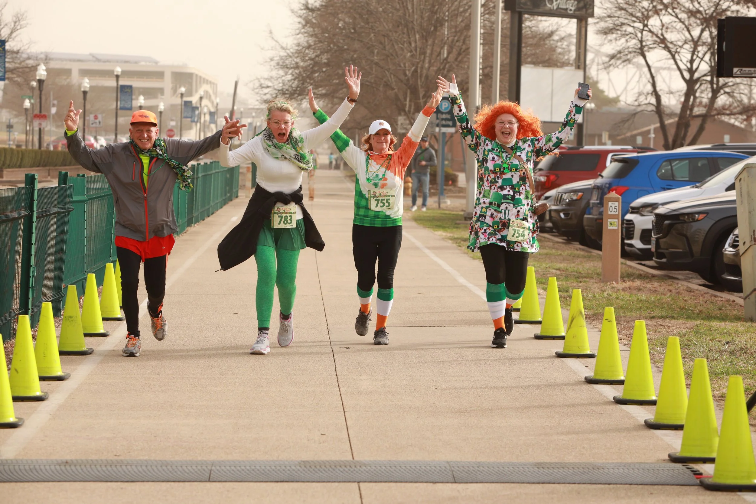 Four people running together in a race, raising their arms in celebration or cheer, on a paved sidewalk lined with yellow cones, with parked cars and trees in the background.