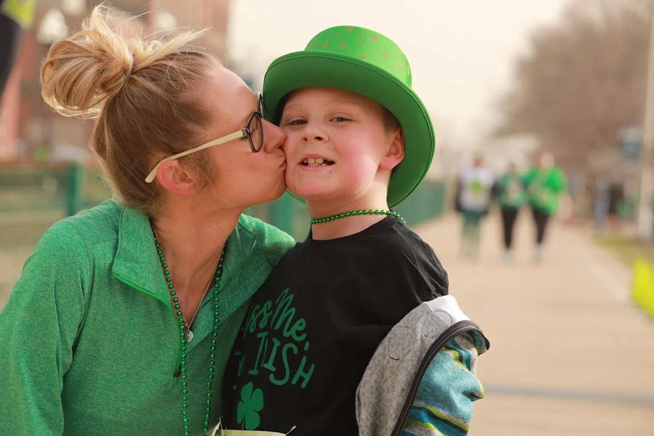 A woman in glasses and a green jacket kisses a young boy wearing a green St. Patrick's Day hat and beads at an outdoor event.
