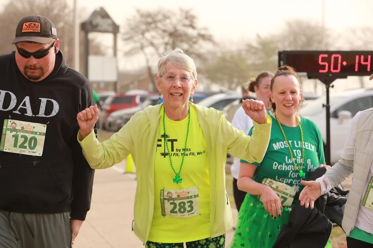 A group of people participating in a race, with a woman in the center raising her fists and smiling. She wears a yellow jacket, a bright yellow shirt, and a green beaded necklace. Other participants are visible around her, and a race timer reads "50: